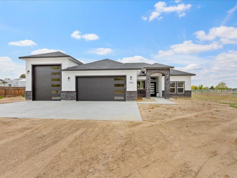 A large white house with a black garage door is sitting on top of a dirt field.