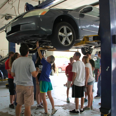 Teens gathered around a car lifted in a shop, observing a mechanic.