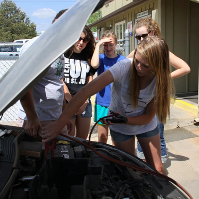Teens gathered around a car with an open hood, jumper cables connected to the battery.