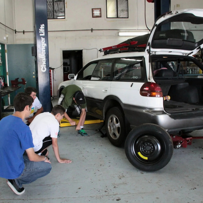 Teens changing a car tire in a garage, a white station wagon on a lift, spare tire on the floor.