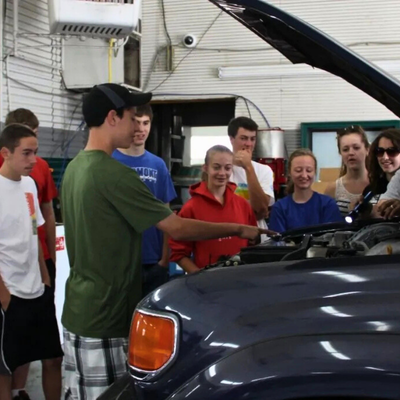 Teenager pointing at car engine, with a group of teens surrounding vehicle.