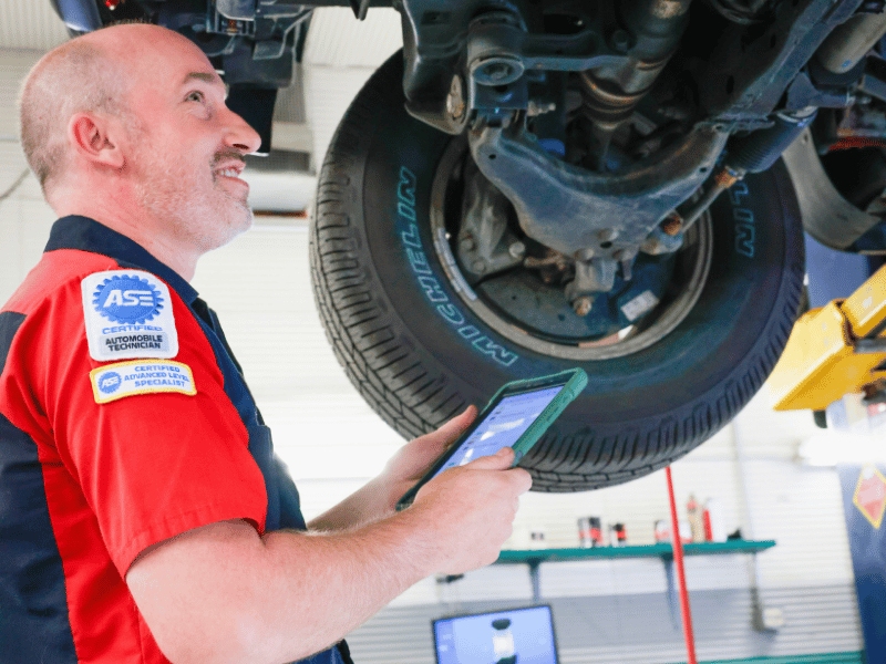 Eric inspecting vehicle's undercarriage with a tablet. Smiling, in red shirt, blue and gray garage, tire visible.
