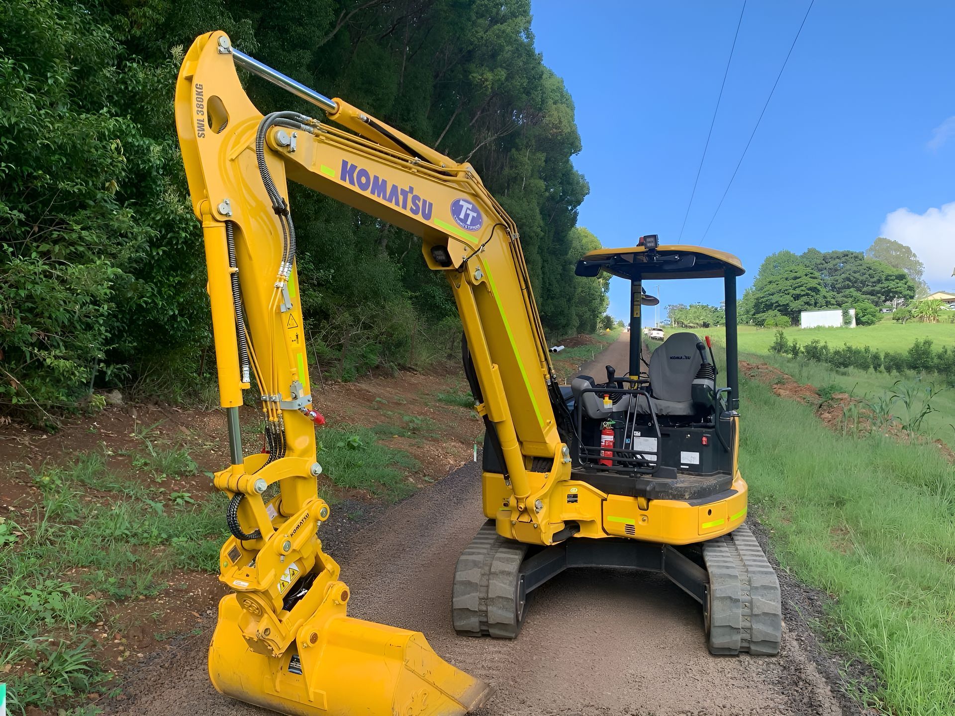 Yellow Komatsu excavator on a dirt path next to greenery under a bright blue sky.