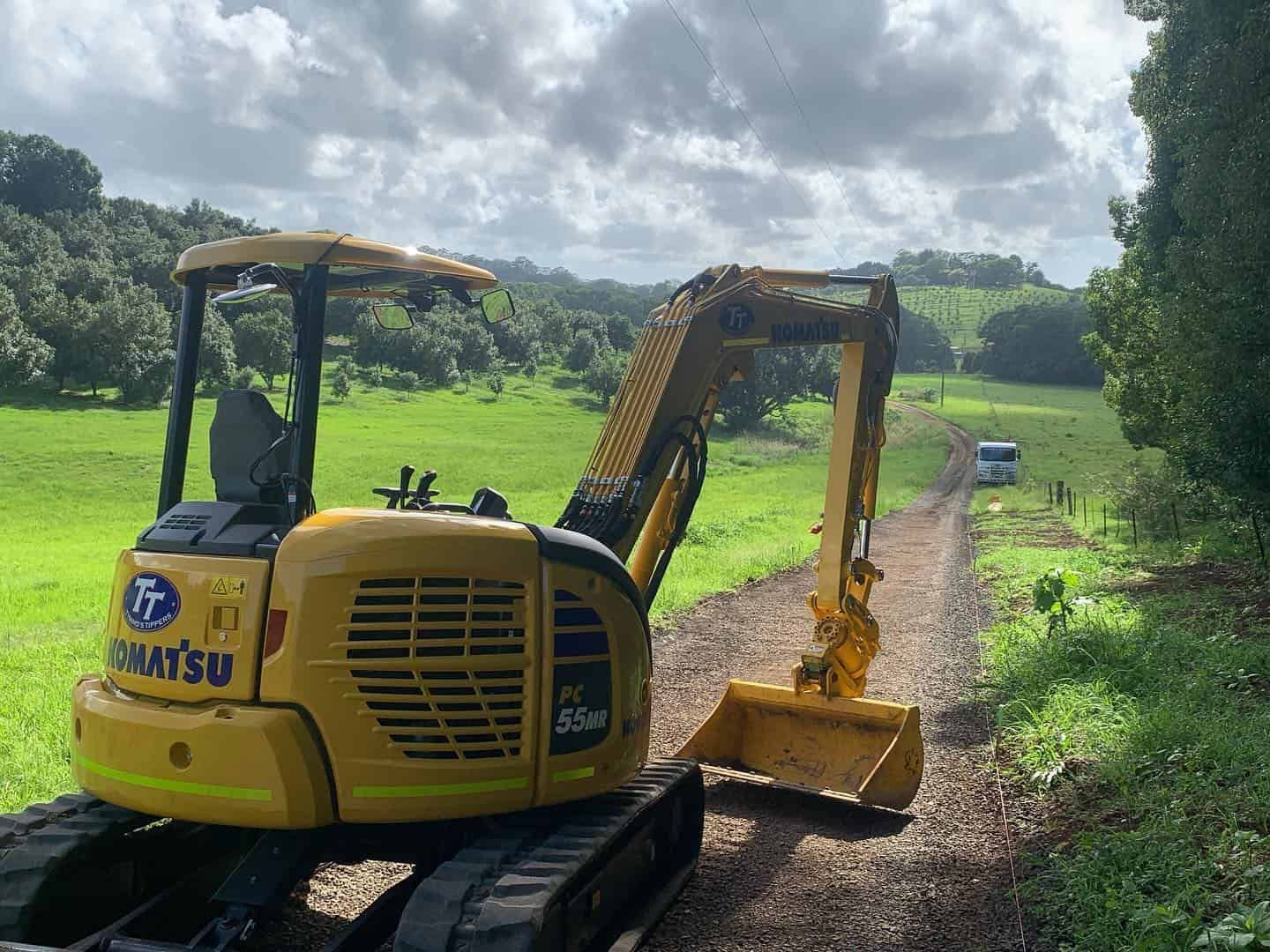 A Yellow Excavator Is Driving Down A Dirt Road In A Field — Tymmo's Tippers In Alstonville, NSW