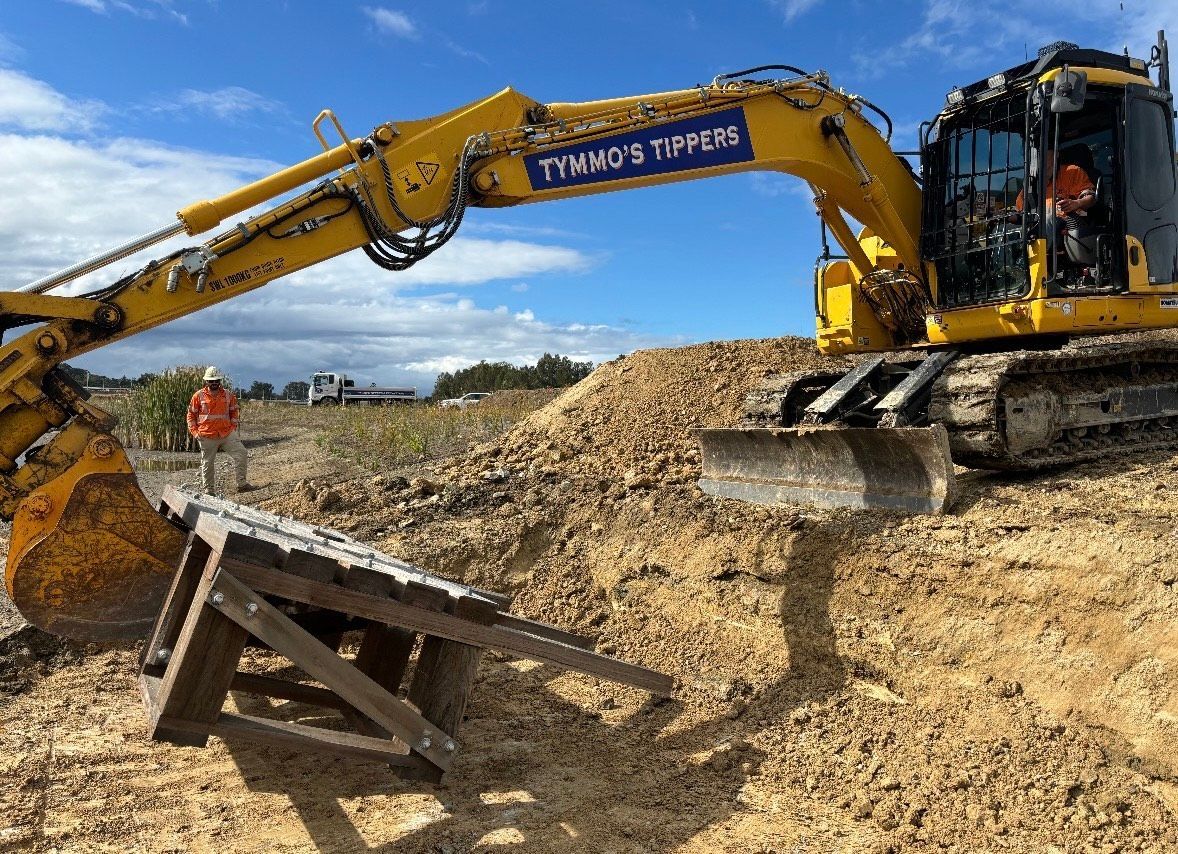 Yellow excavator digging earth on a construction site. A person in an orange vest stands nearby.