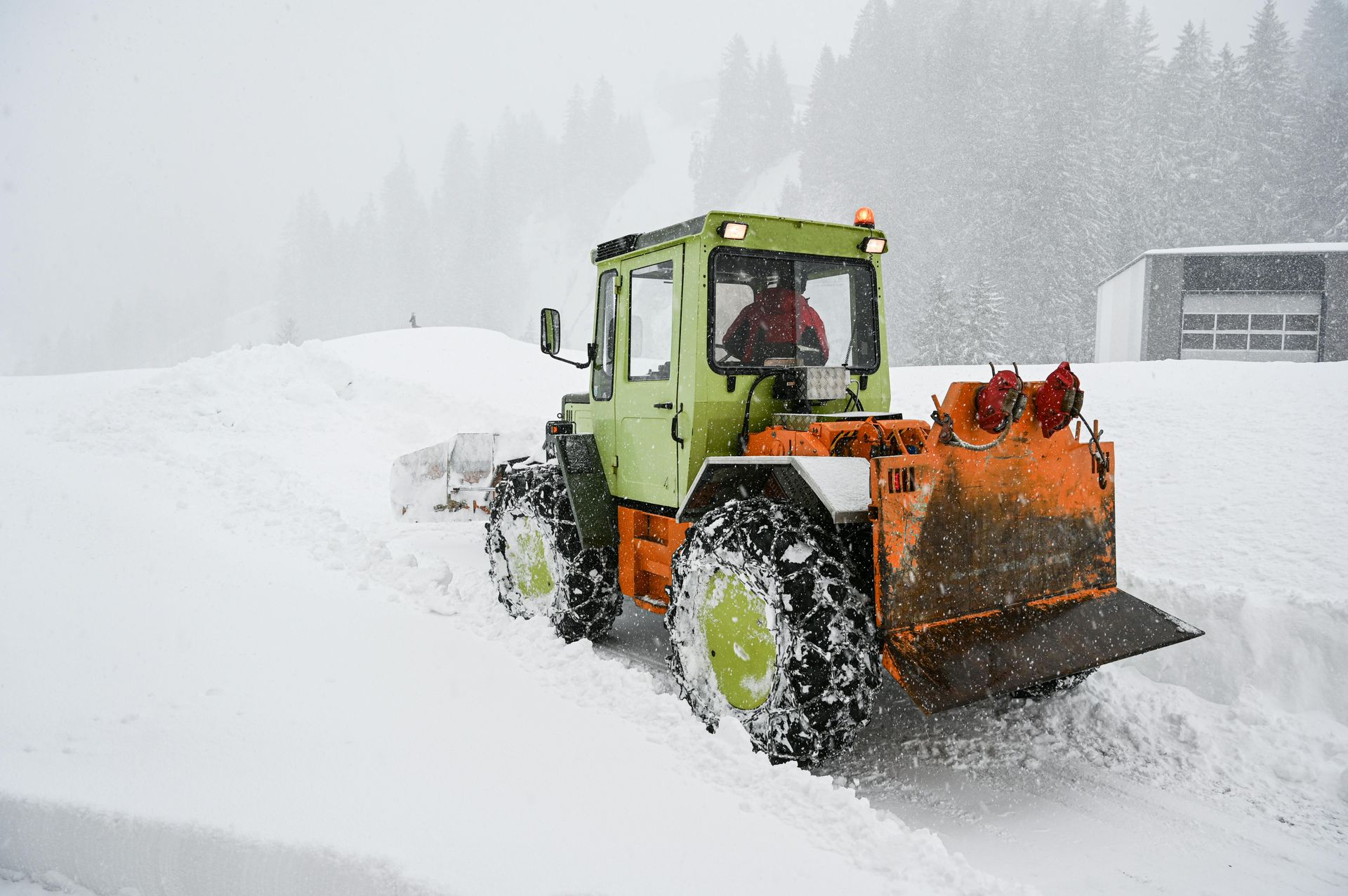 Snowplow clearing snow on a snowy day. Green and orange tractor with the driver inside.