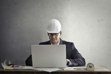 Man in suit and hard hat works on a laptop at a desk with blueprints and tools.