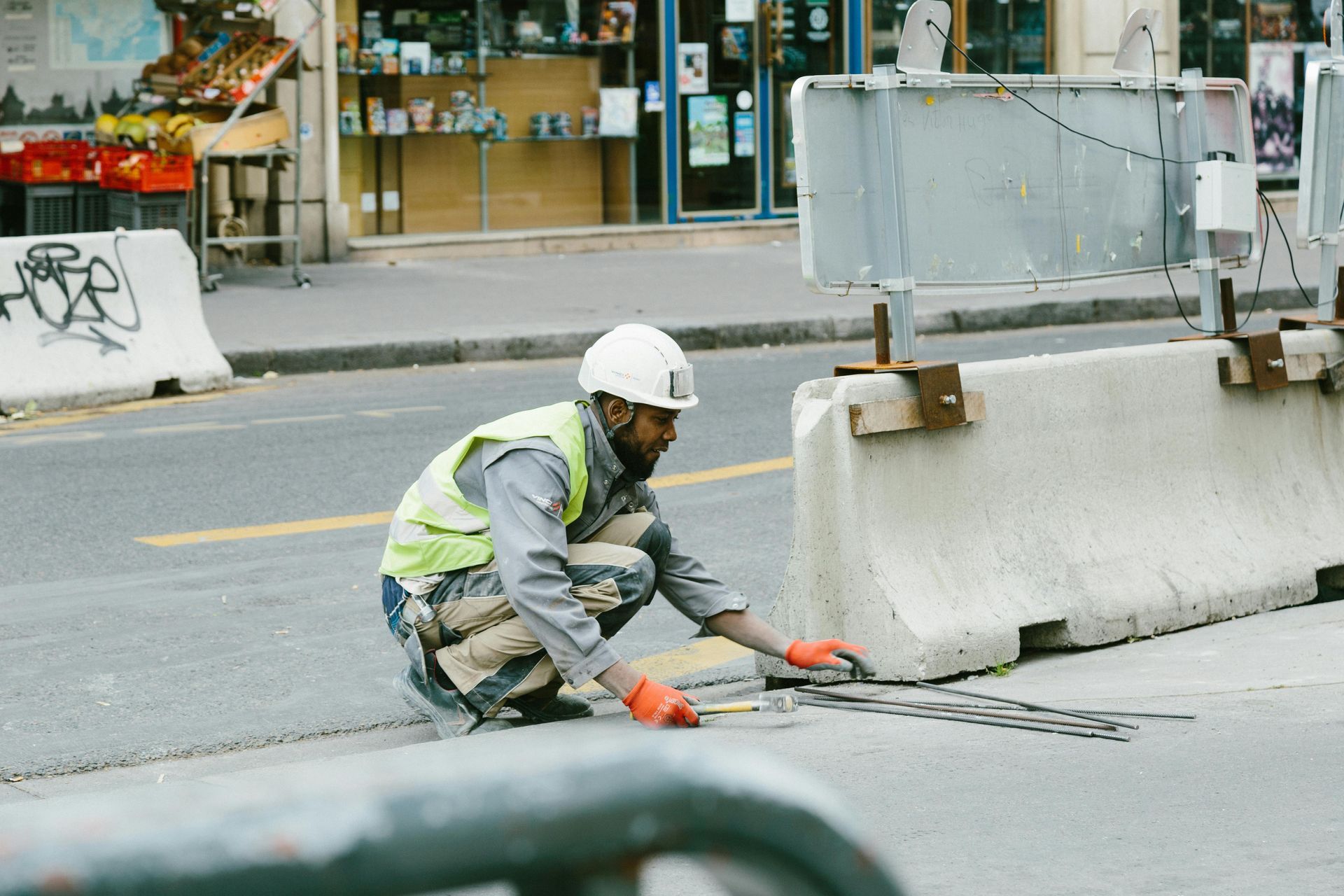 Construction worker in white helmet and orange gloves works on road near concrete barriers.