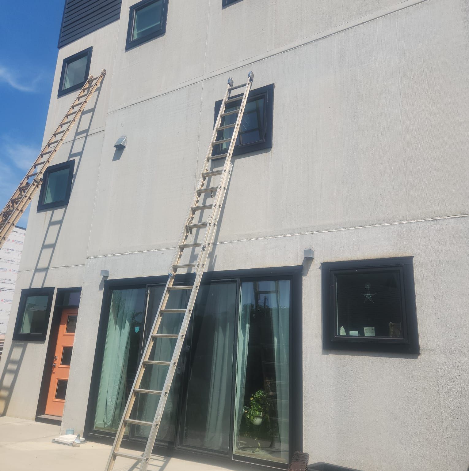 Three-story building exterior with ladders leaning against the stucco wall; windows and doors are dark-framed.