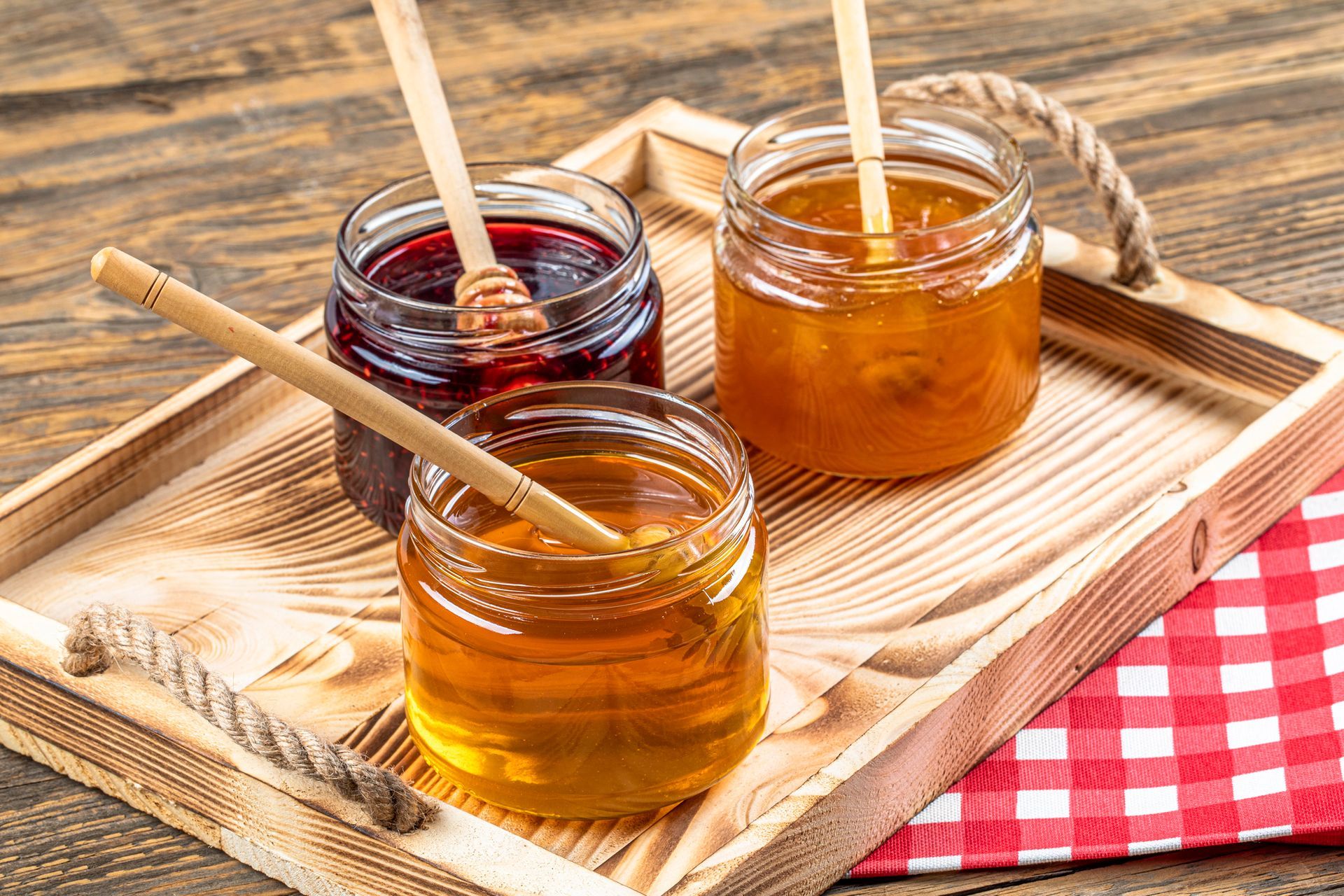Tre barattoli di miele e marmellata con cucchiaini su un vassoio di legno, con un panno a quadretti rossi e bianchi.