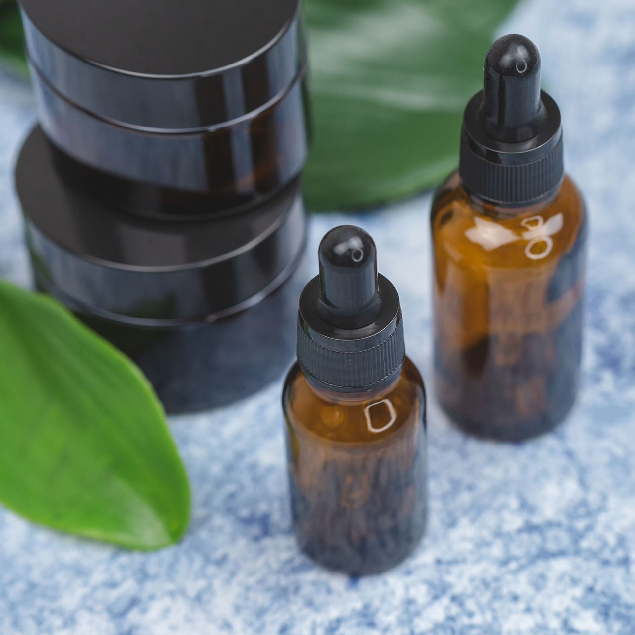 Flaconi contagocce in vetro ambrato e barattoli cosmetici neri su sfondo blu, accanto a una foglia verde.