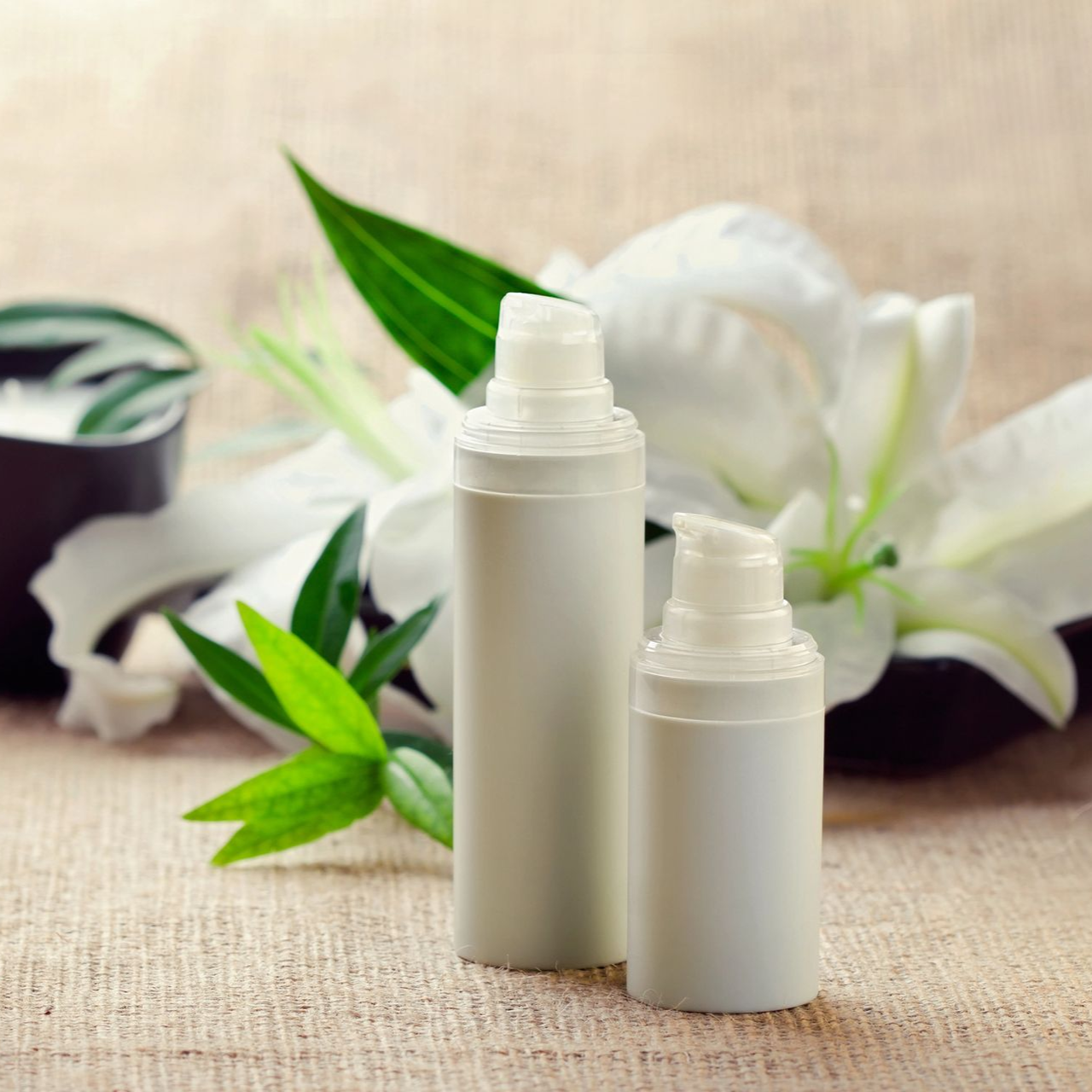 Due flaconi bianchi di prodotti per la cura della pelle con erogatori a pompa, fiori bianchi e foglie verdi.