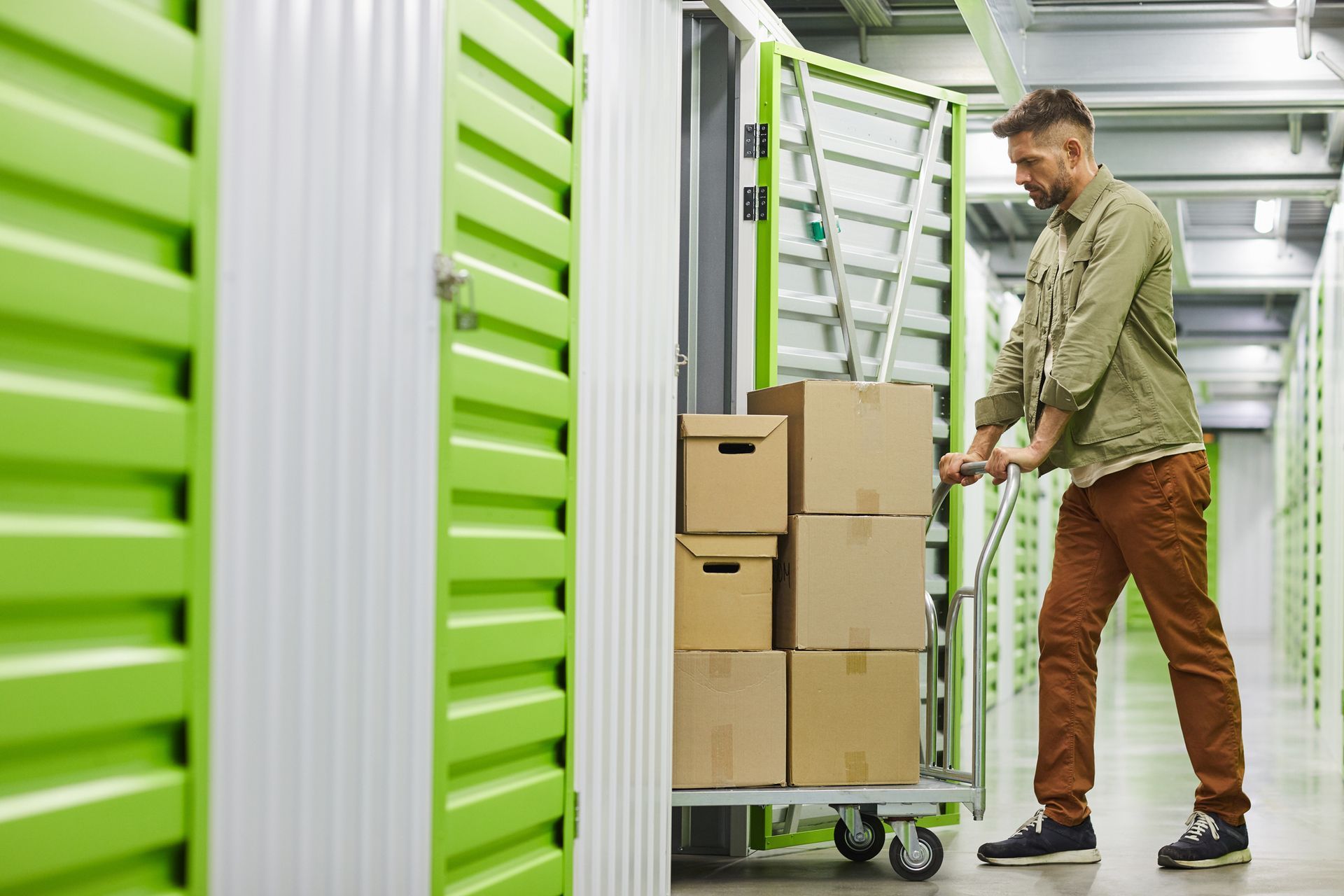 Side view of a man loading a cart with cardboard boxes into a Self Storage unit with green doors.