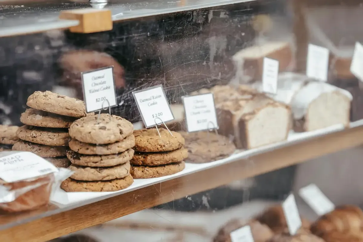 ServSafe Food Handler with a glass bakery case displaying stacks of chocolate chip and oatmeal raisin cookies.