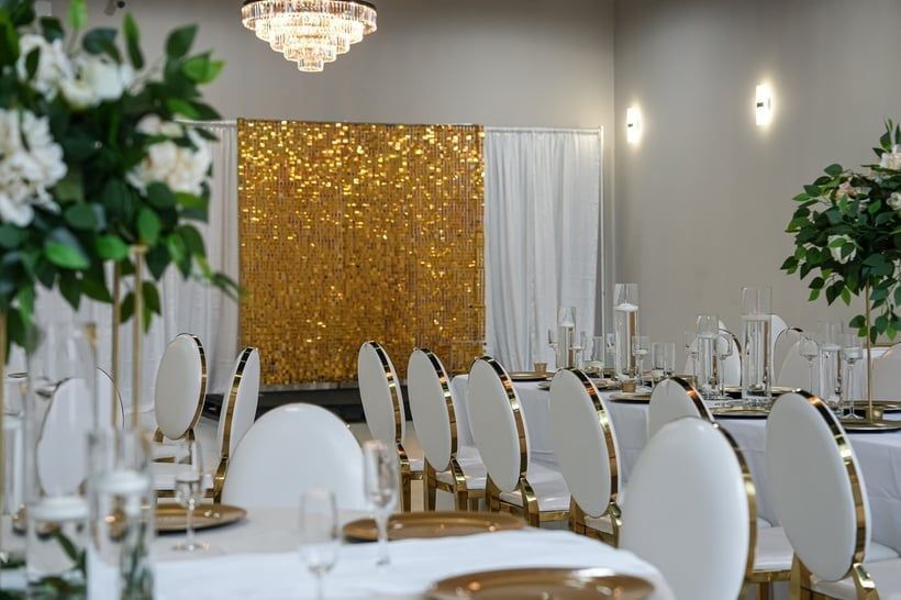 Elegant banquet hall with white chairs, gold table settings, chandeliers, and a gold sequin backdrop
