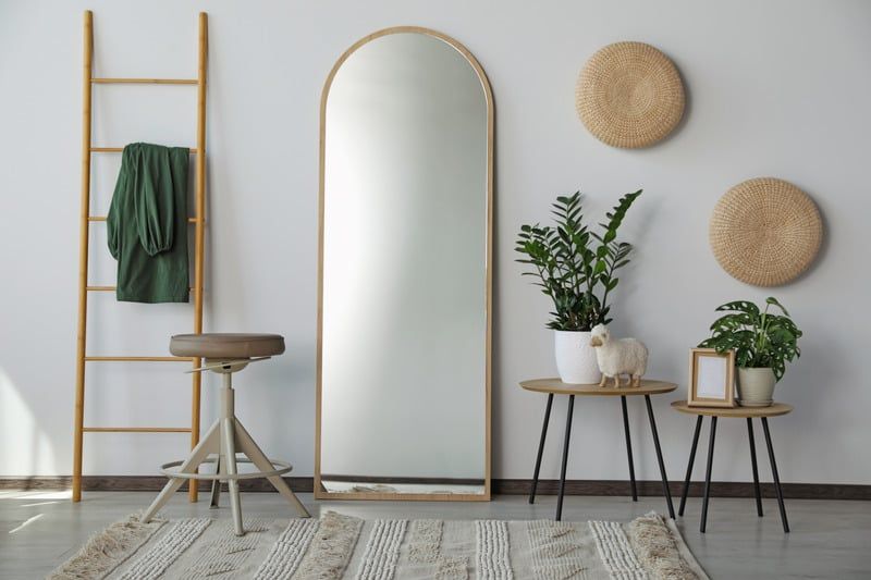 Minimal living room with arched mirror, ladder, stools, plants, and woven wall decor on a light rug.