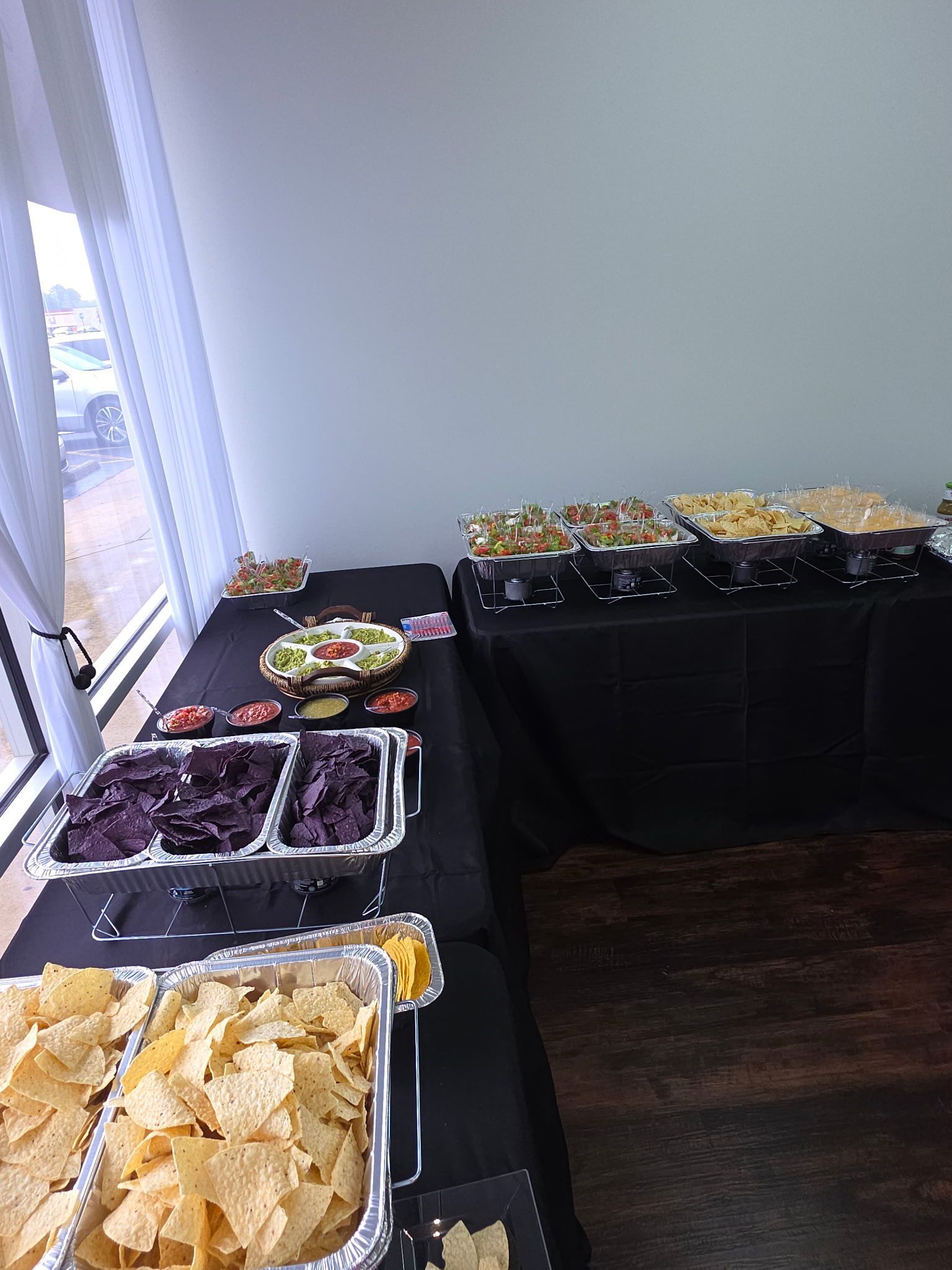Buffet table with trays of chips, grapes, and salads set up beside large windows
