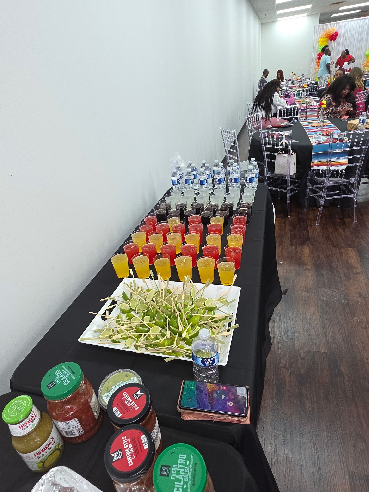 Table with assorted drinks, jars, and trays of food along a hallway beside a wall.