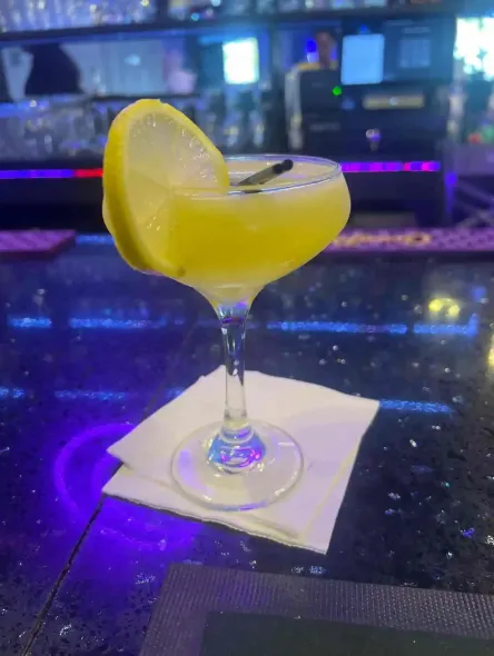 Yellow cocktail in a stemmed glass with a lemon slice, sitting on a napkin at a dimly lit bar.