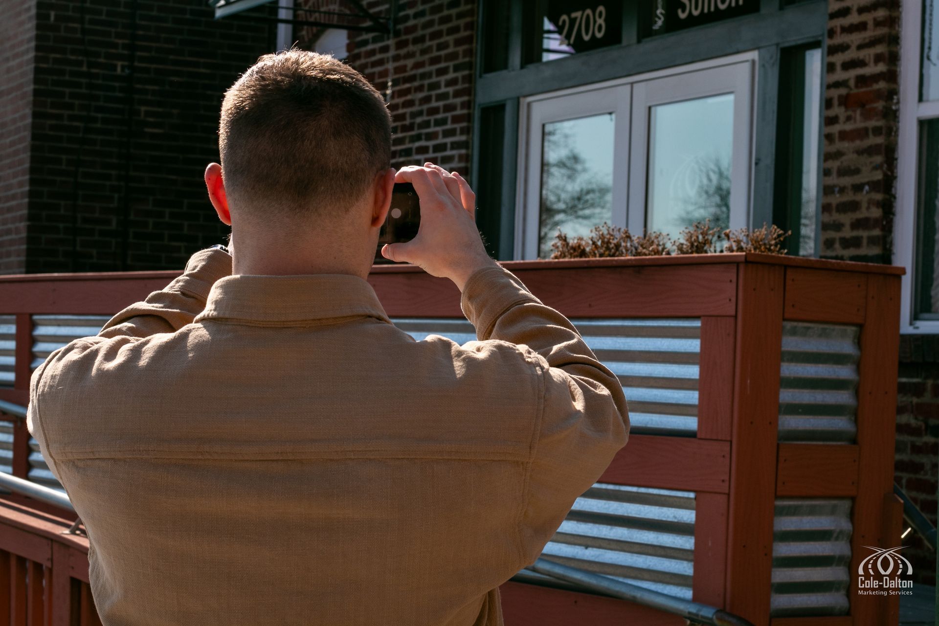 A man is filming a building outdoors. Brown jacket, brick building, sunny.