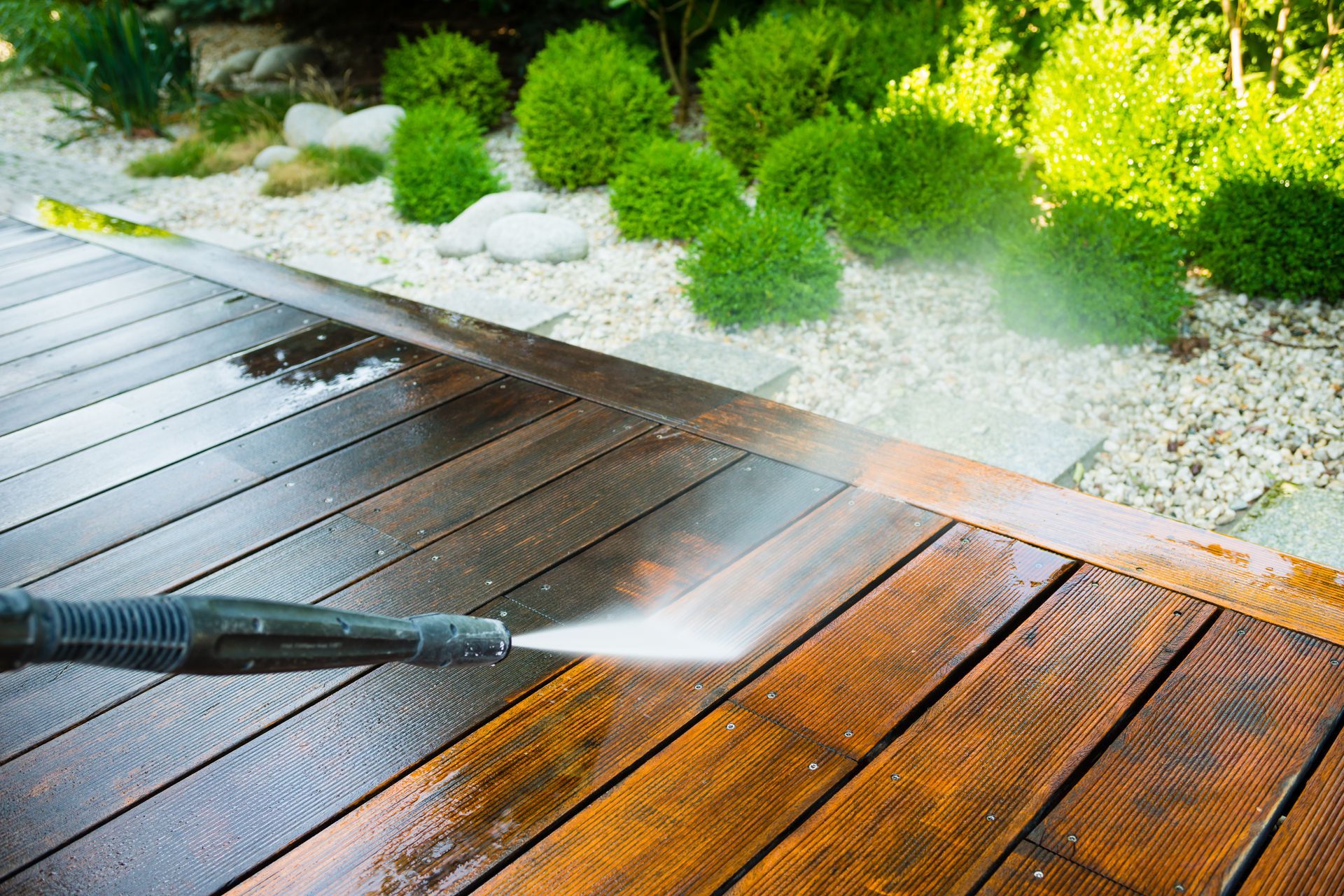 Pressure washing a wooden deck beside a landscaped garden with green shrubs and white gravel