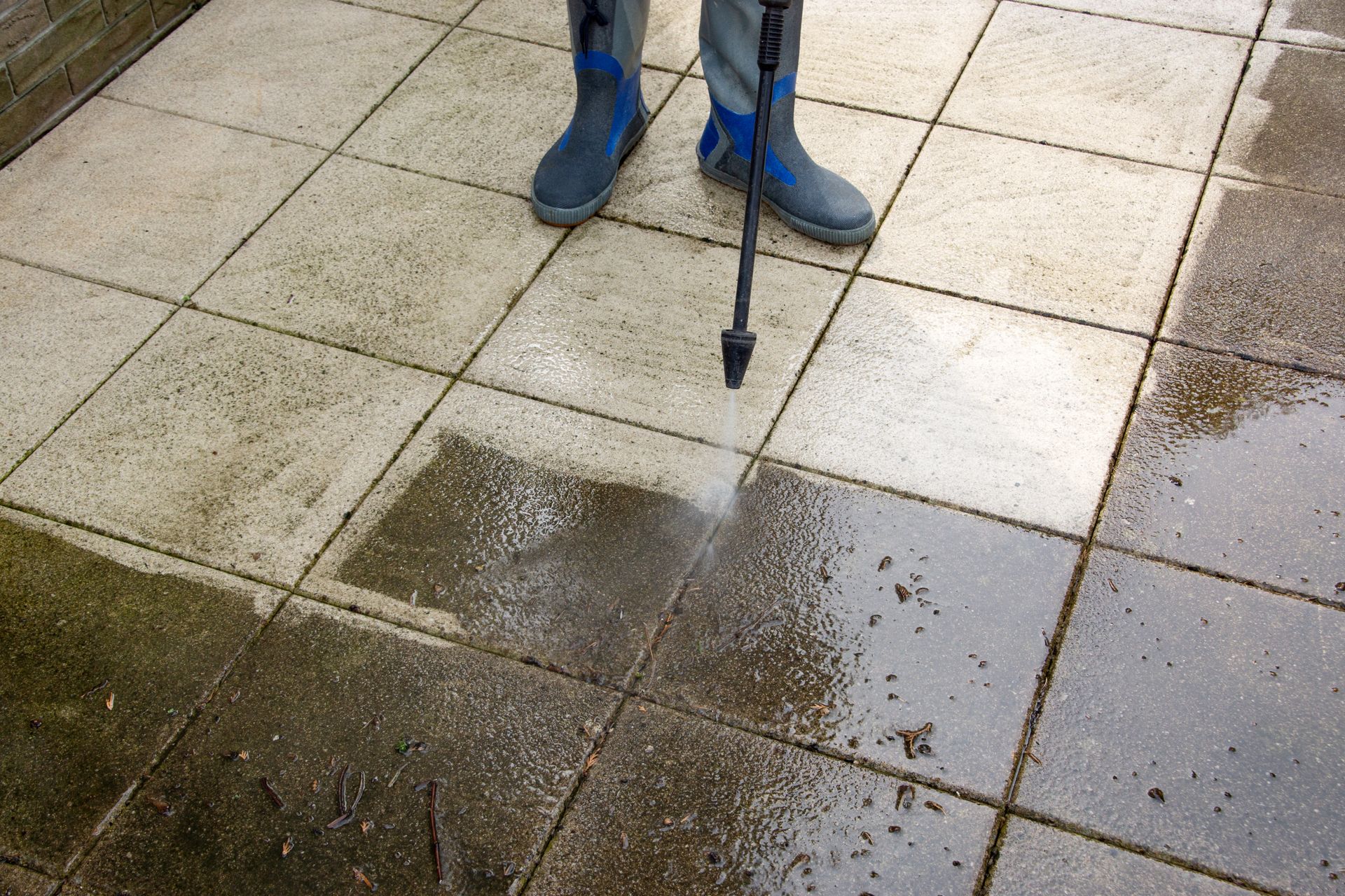 Person using a pressure washer to clean a wet stone patio, with blue boots visible.
