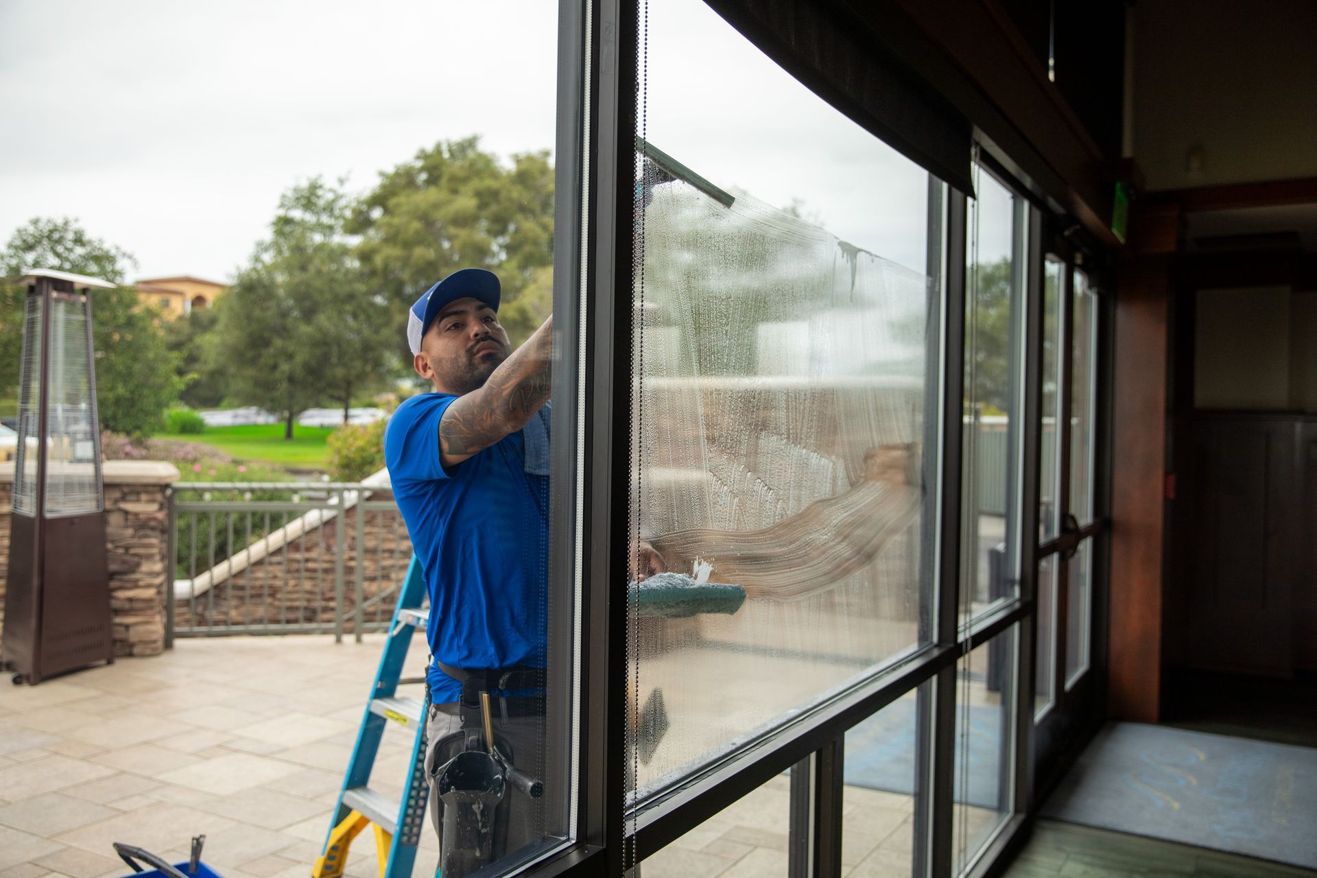 Worker cleaning a large glass window beside a ladder on a rainy day
