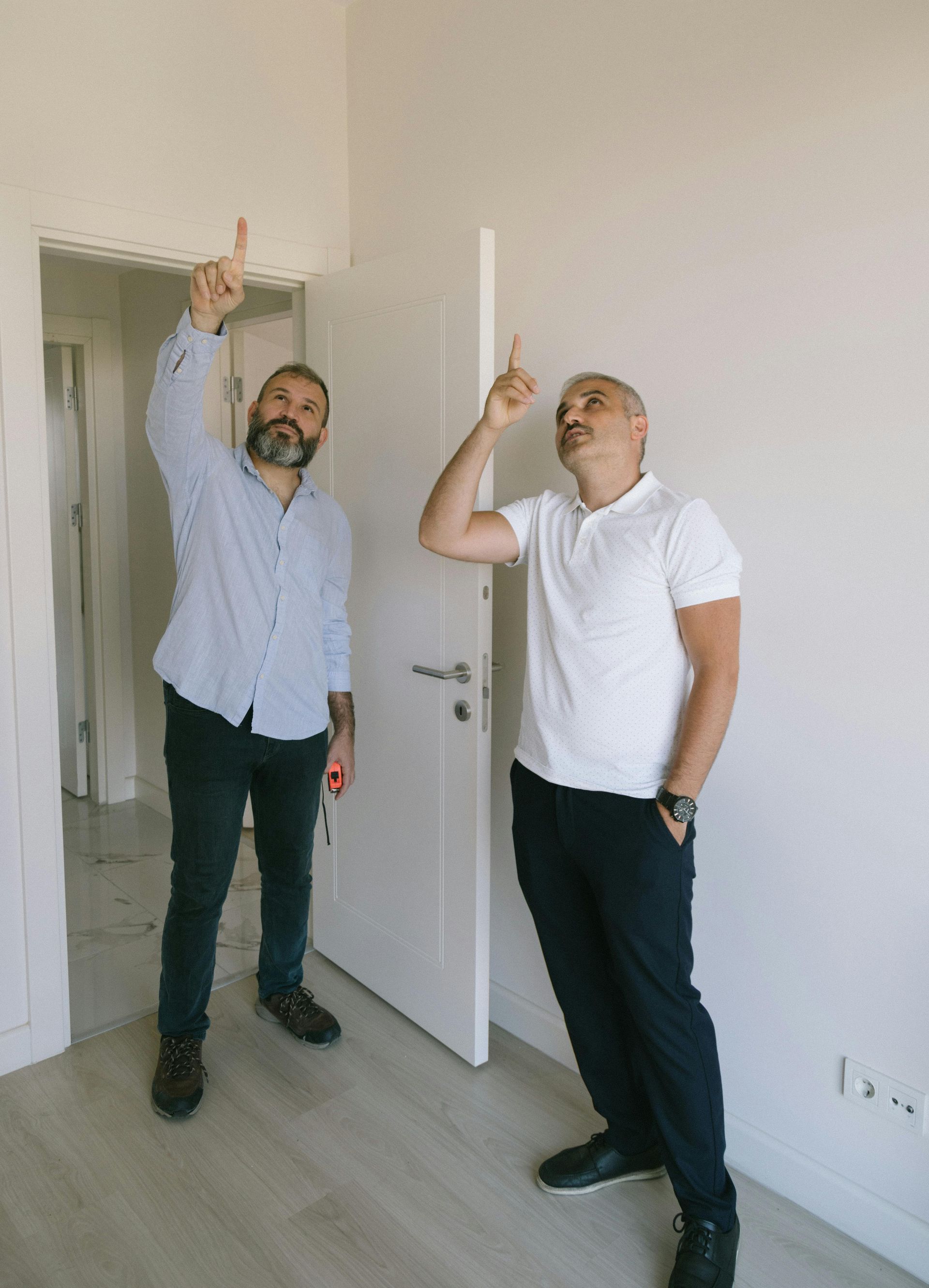 Two men stand in a white-walled room, looking up and pointing at the ceiling while talking.