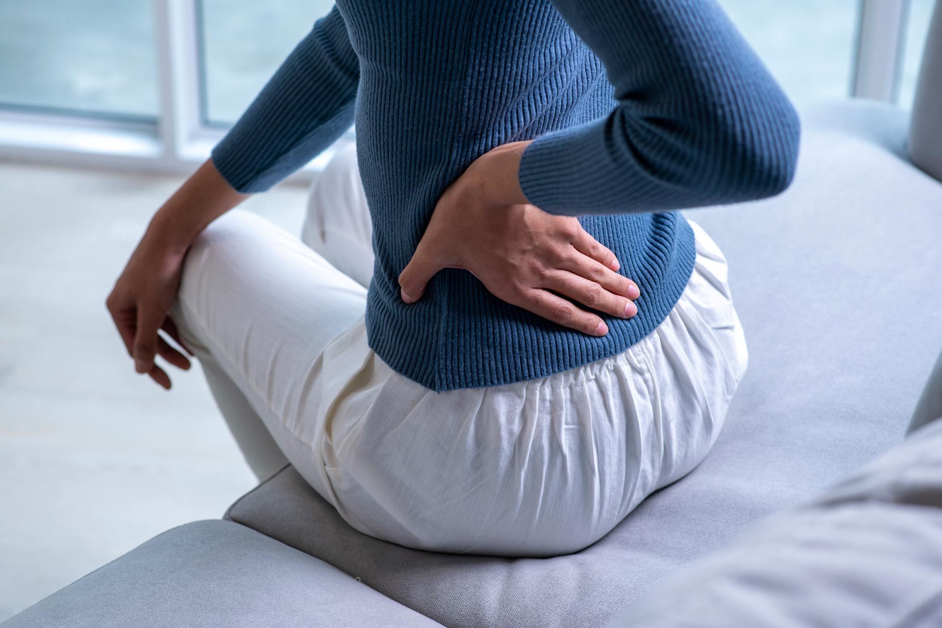 A woman is sitting on a couch holding her back in pain.