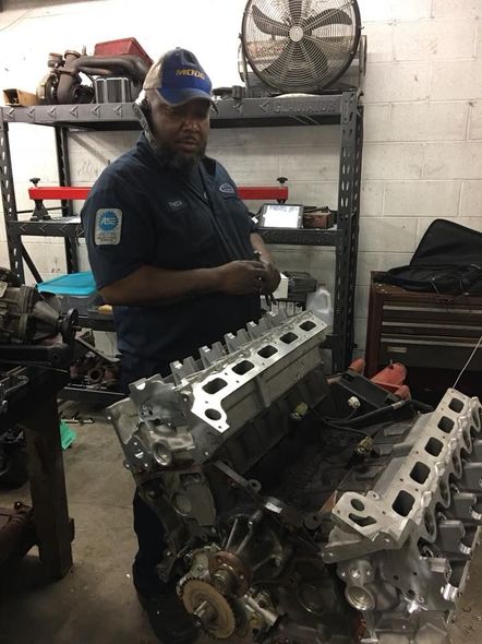 Mechanic holding engine head in a garage; wears blue work uniform and cap.  A dark-skinned man; the setting is cluttered.