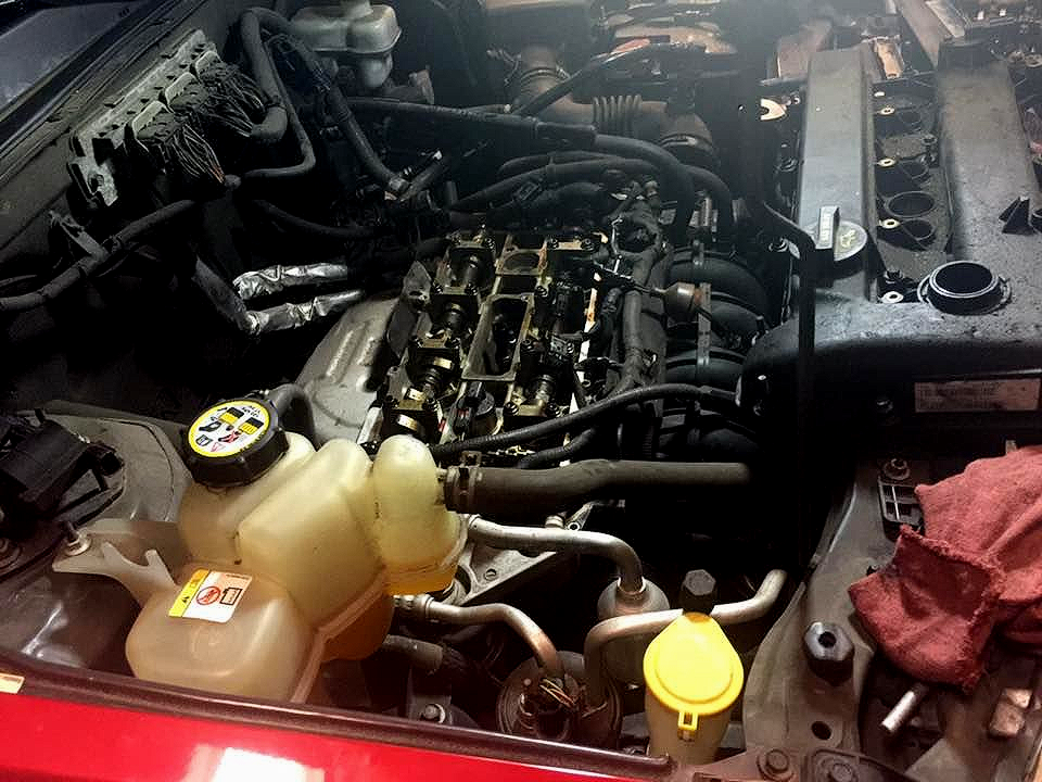 Car engine bay, partially disassembled, showing exposed engine components. Coolant reservoir is visible.