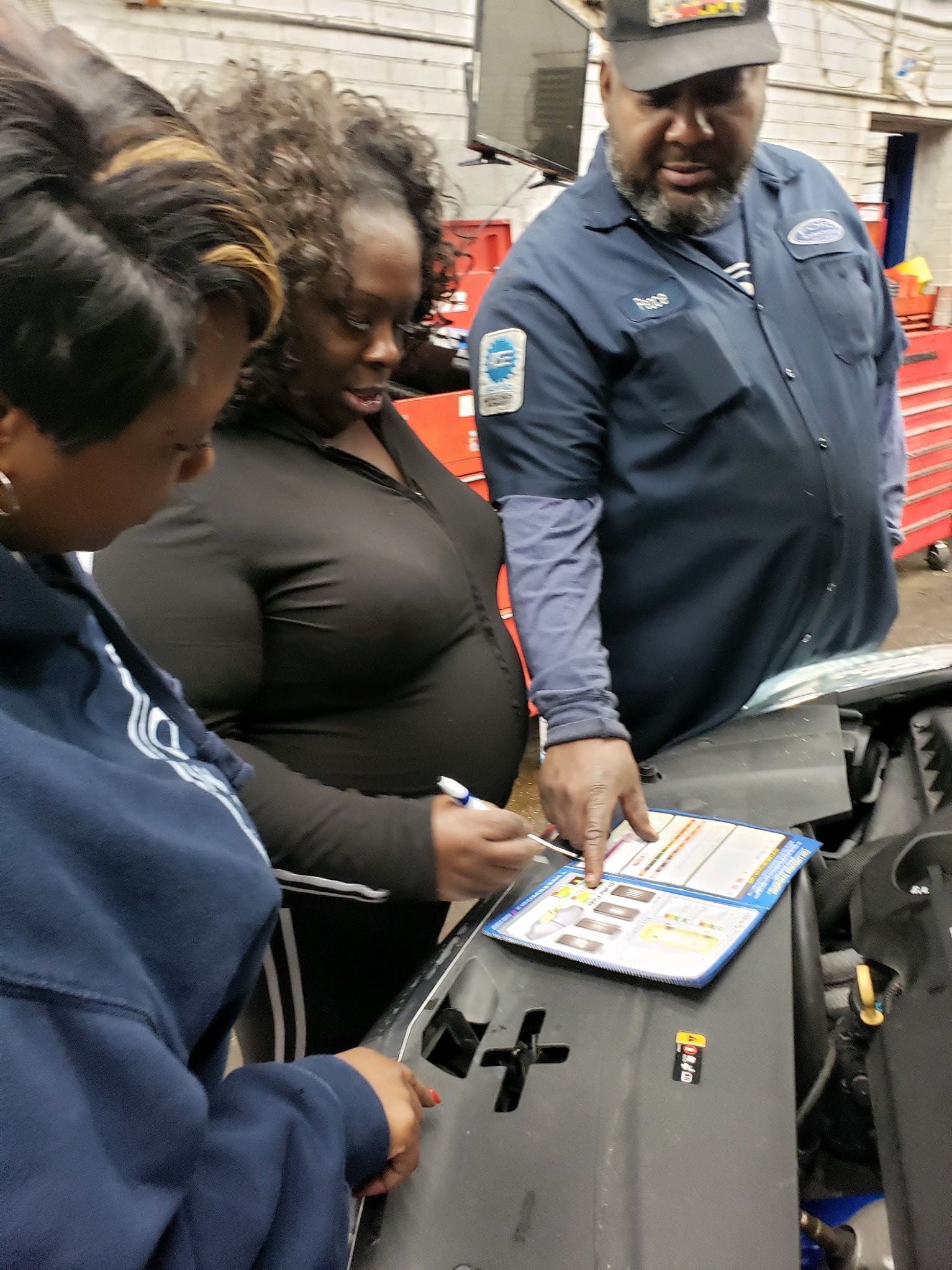 Three women and a mechanic looking at car engine parts, discussing a checklist.