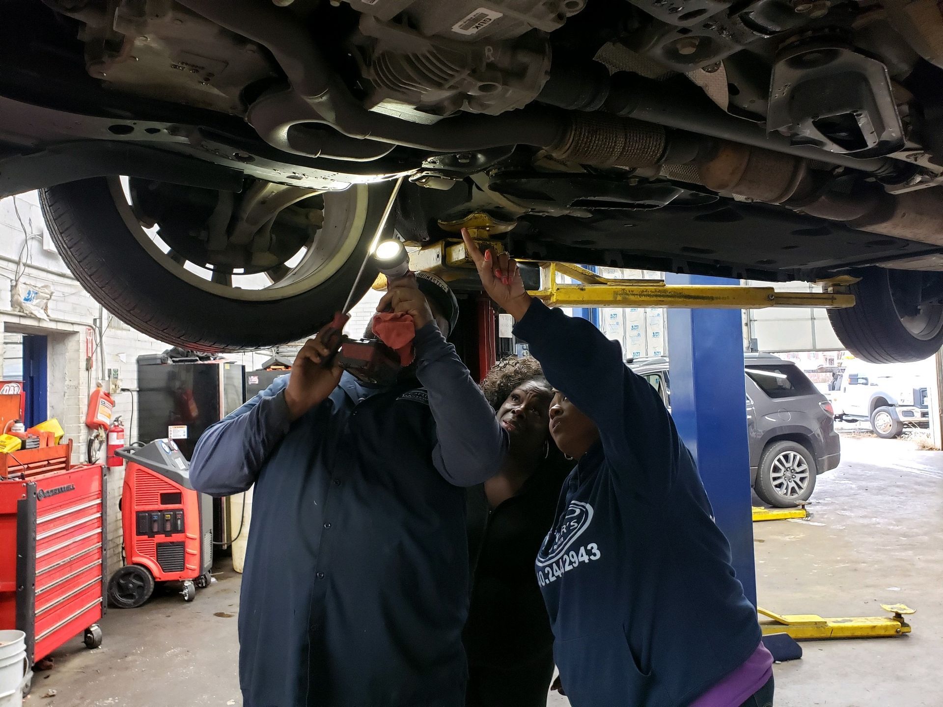 Three people examining underside of a car on a lift in an auto repair shop.