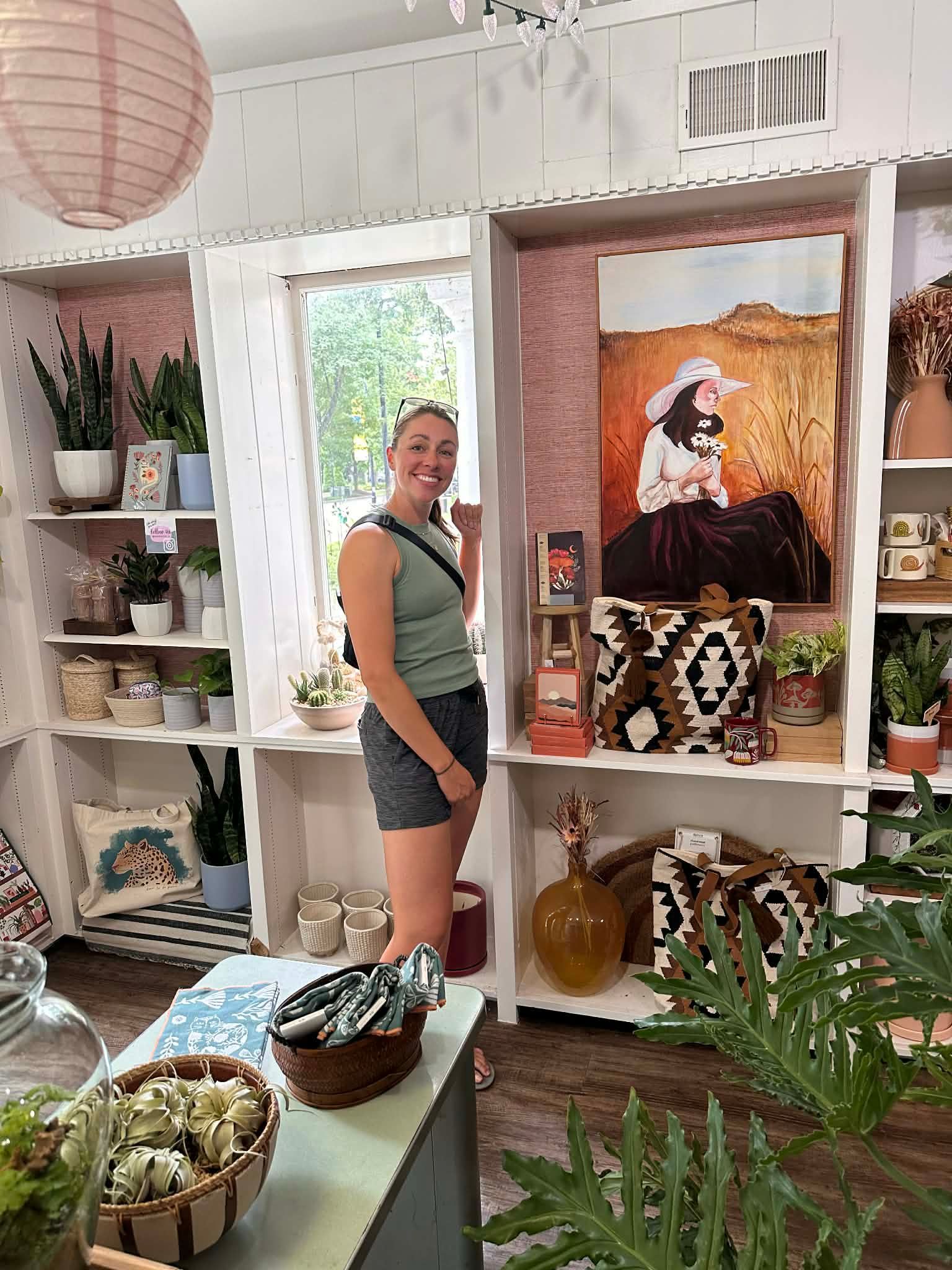 A person stands in a brightly lit boutique shop, surrounded by shelves featuring decor, house plants, and art.