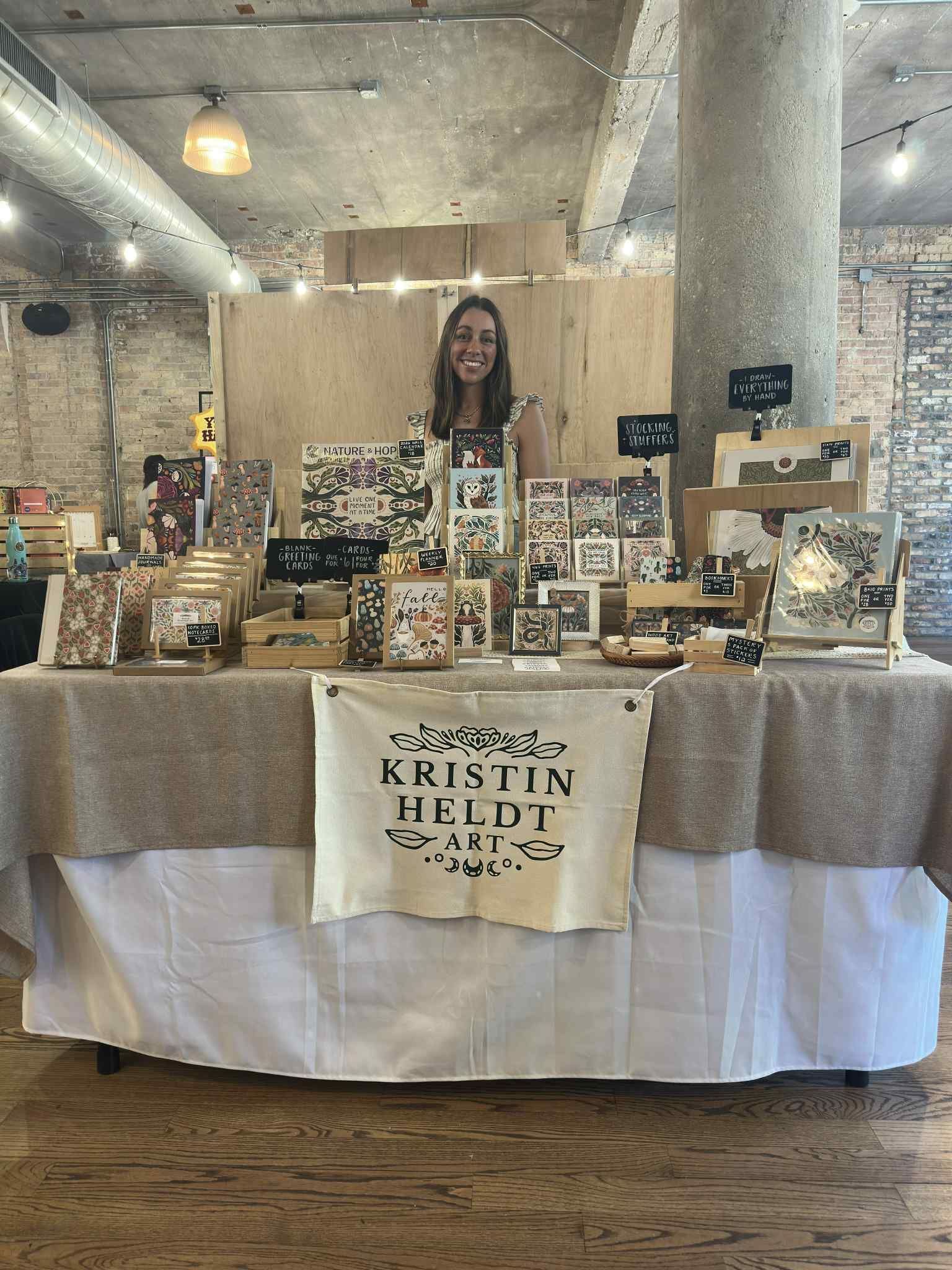 A smiling vendor stands behind a table displaying art prints and goods at a market in a rustic indoor space.