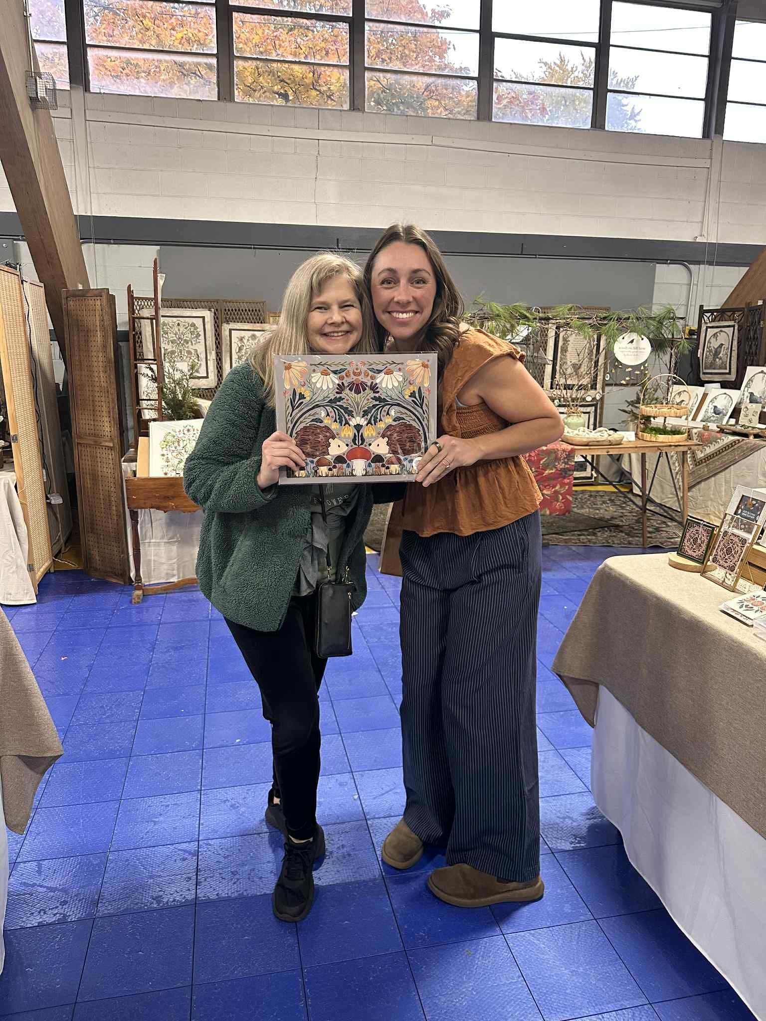 Two people smile while holding a framed artwork at a craft show, set against a backdrop of displays and blue flooring.