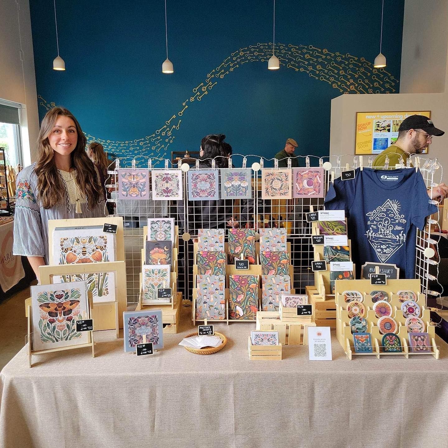 A woman stands behind a market stall table covered in colorful art prints, greeting cards, and shirts in a store.