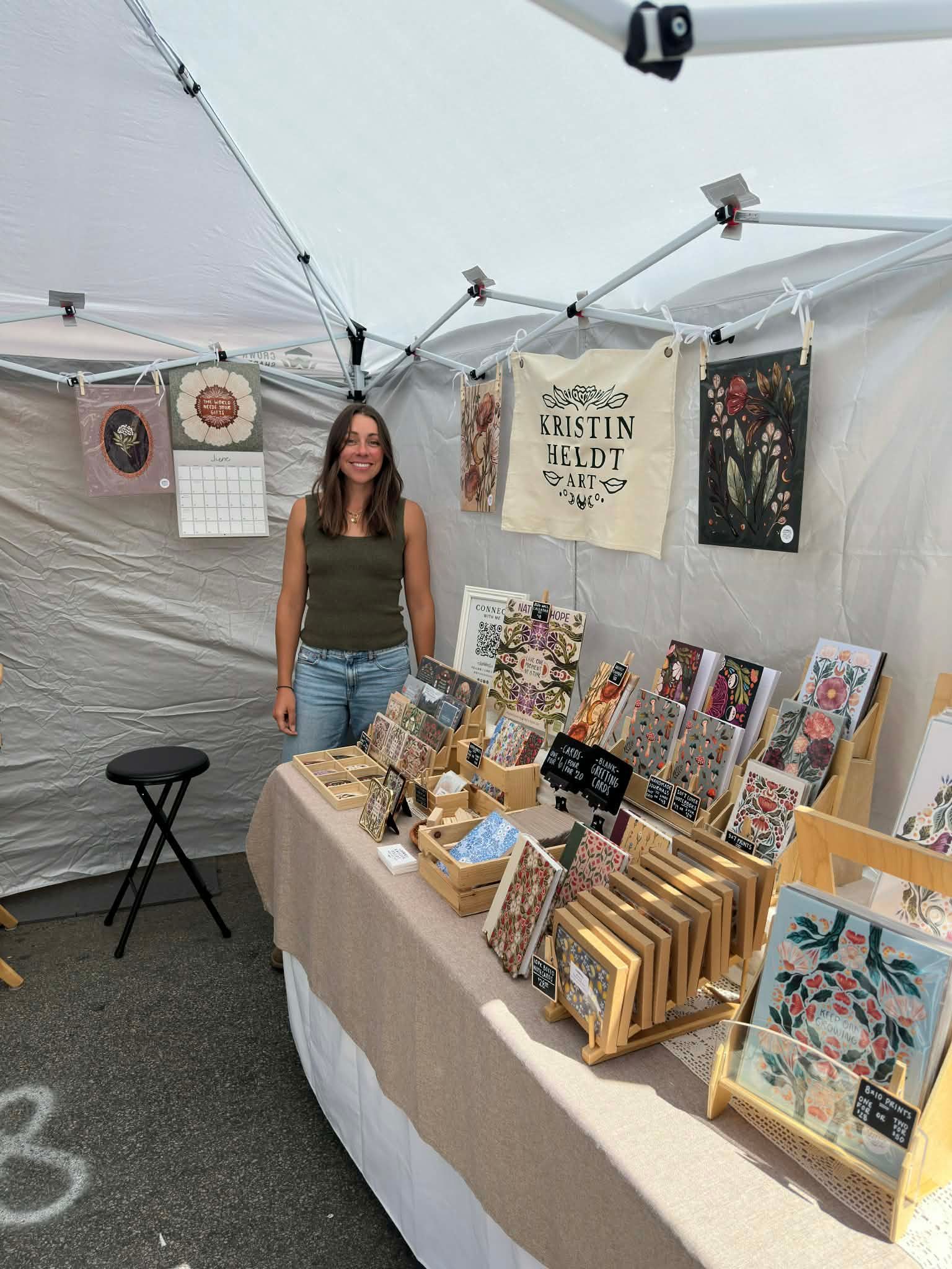 A person stands behind a table at an outdoor market stall, displaying floral and patterned art prints, cards, and decor.