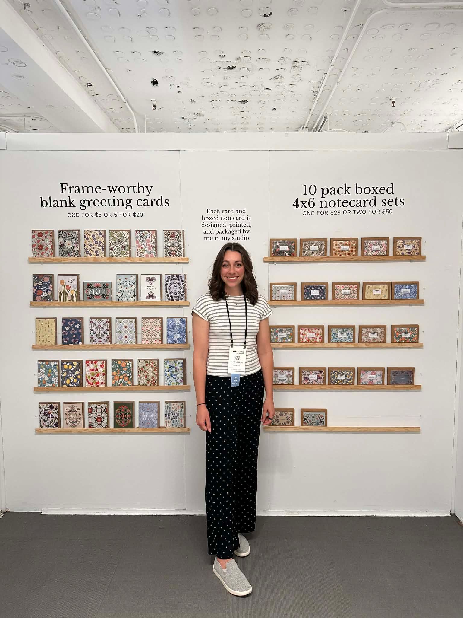 A smiling person stands in a gallery between two walls lined with small, framed square artworks displayed on shelves.