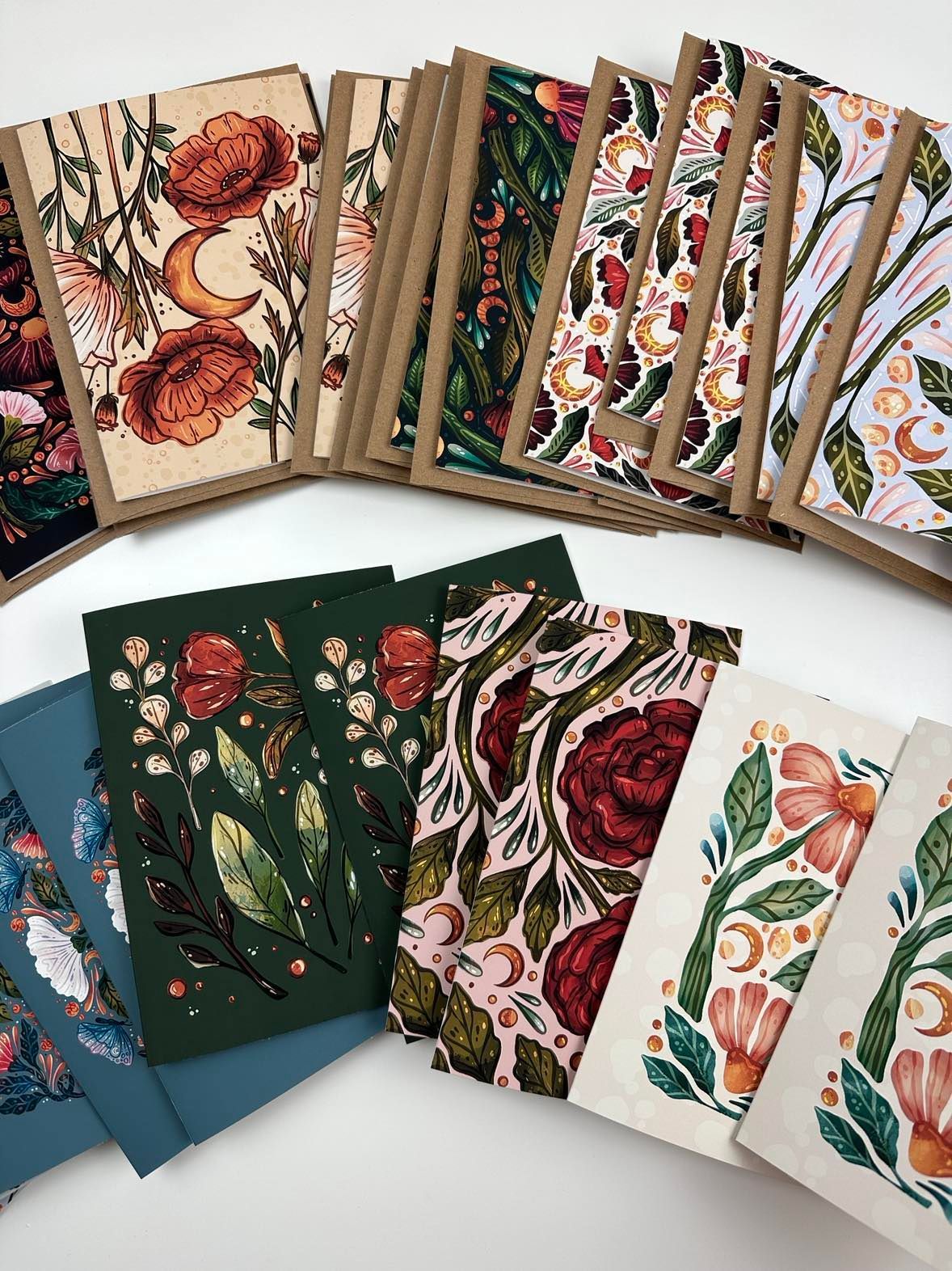 A flat lay of assorted greeting cards featuring botanical and celestial illustrations in floral, dark green, and cream hues.