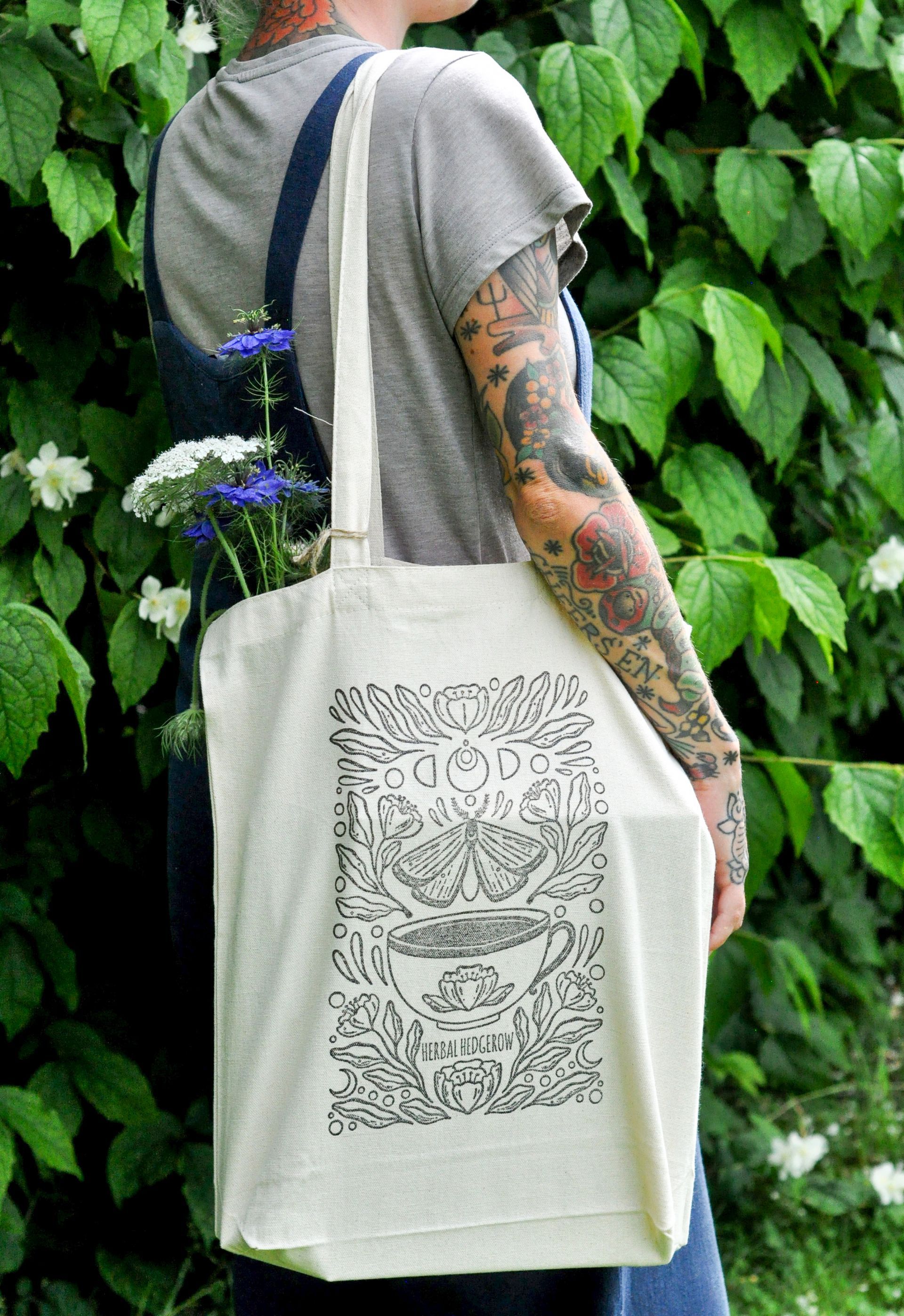 A woman with tattoos is holding a tote bag with a cup of tea on it.