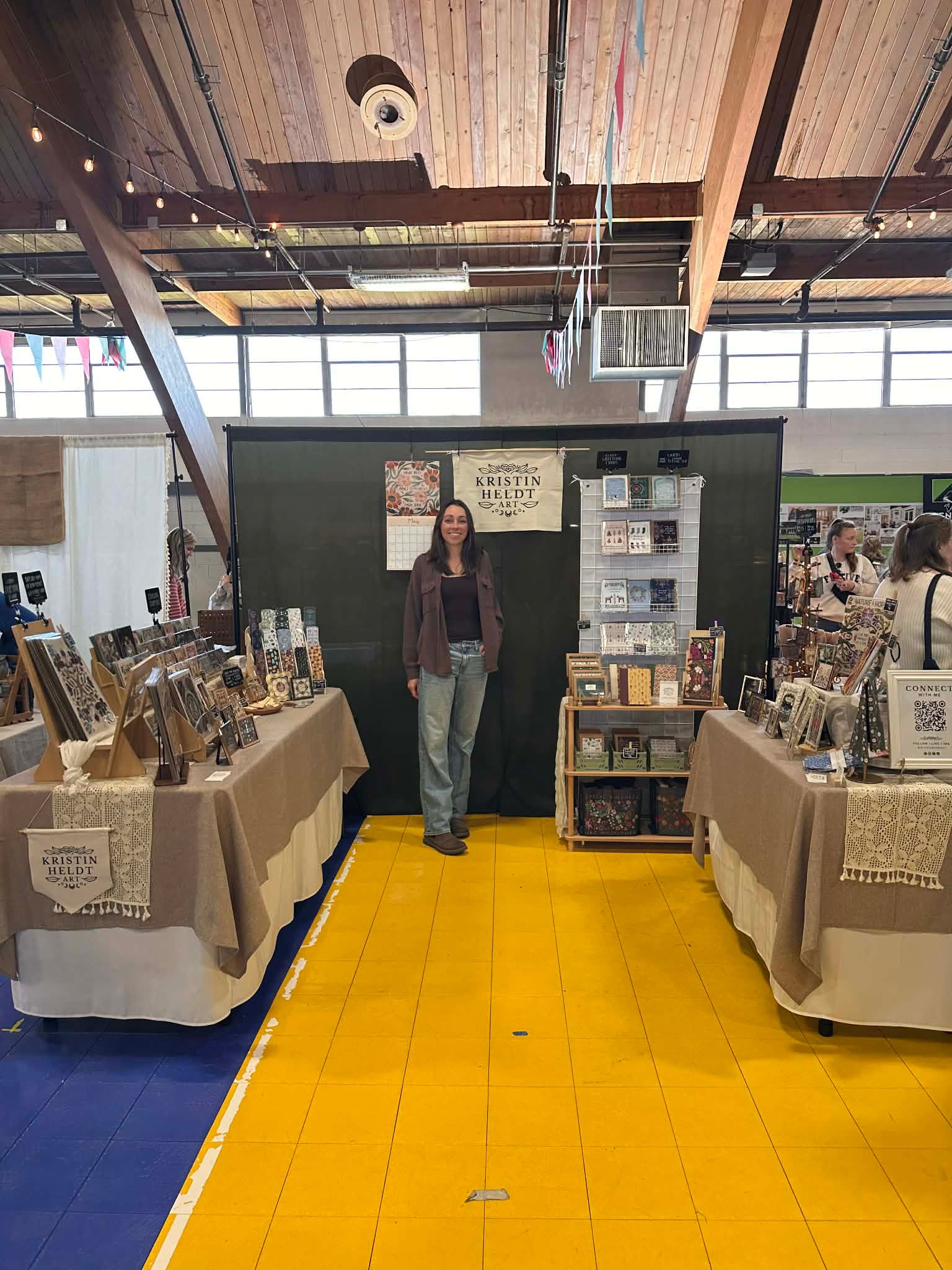 A person stands in a booth at a craft fair, centered between two tables display items on beige cloths over a yellow path.