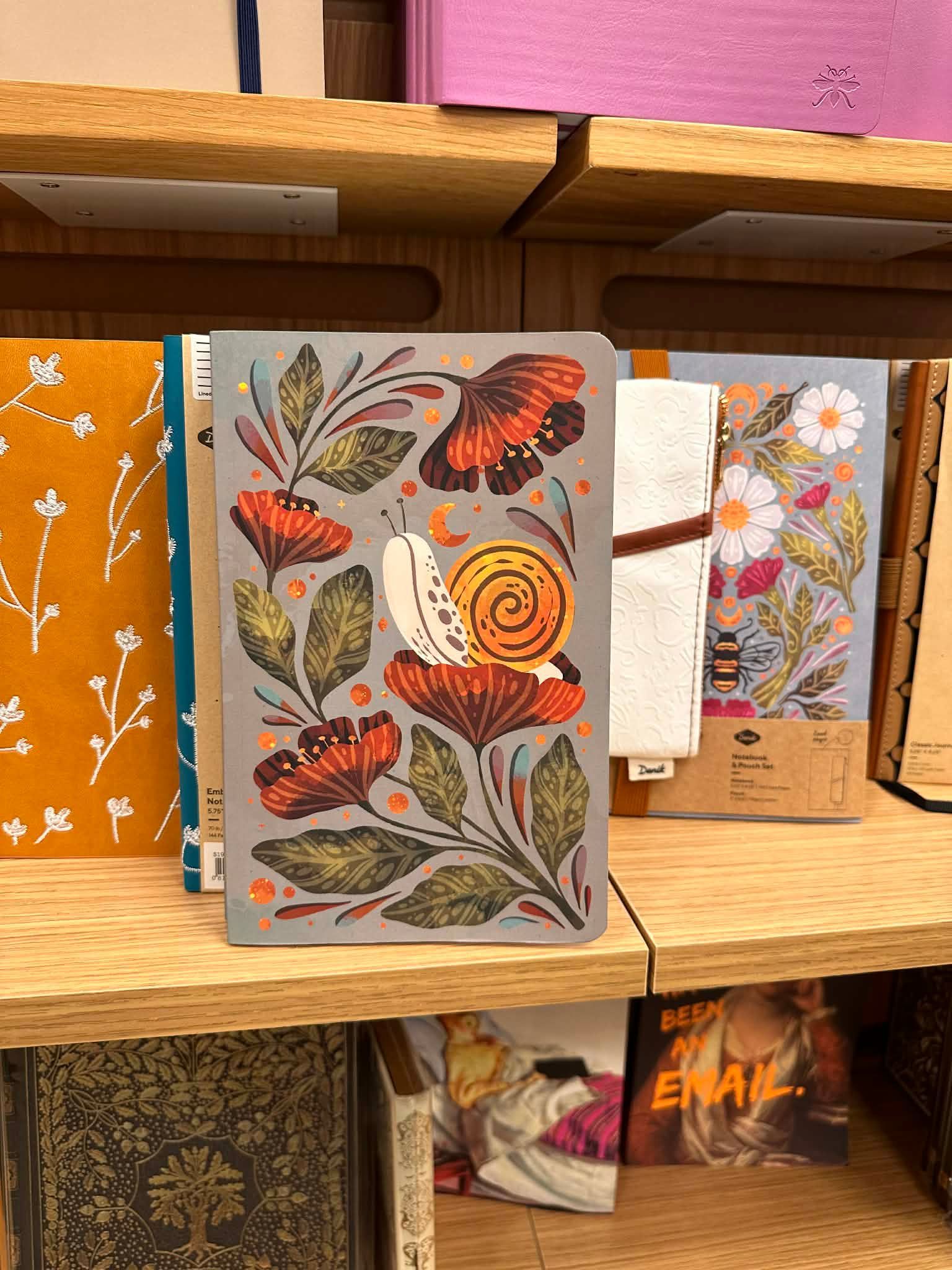 A gray notebook featuring a snail and red flower illustrations on a store shelf surrounded by other journals.