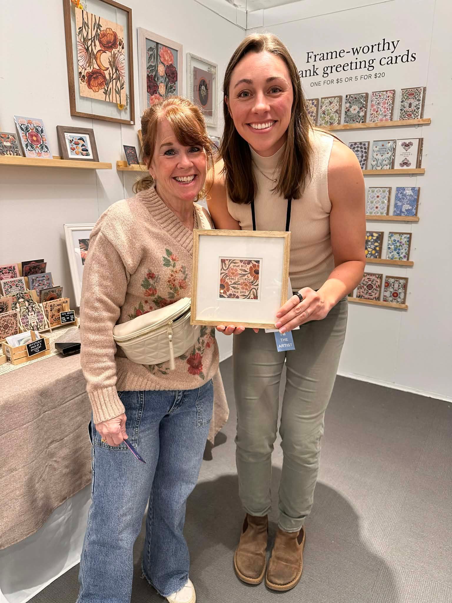 Two people stand in an art booth, smiling while holding a small framed piece of artwork amidst shelves of displayed cards.
