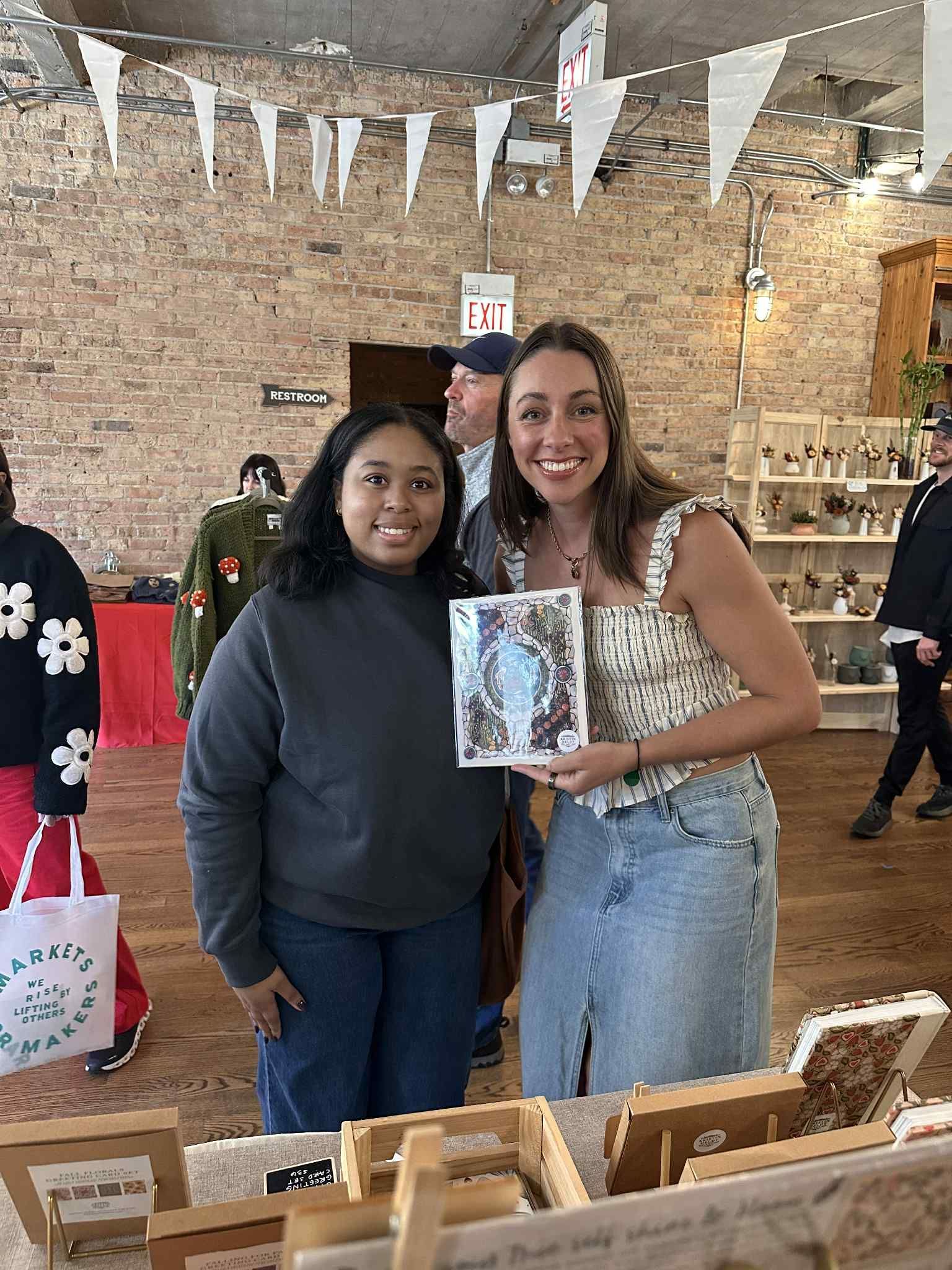 Two people smiling while holding a framed art piece in a rustic shop with brick walls and hanging bunting.