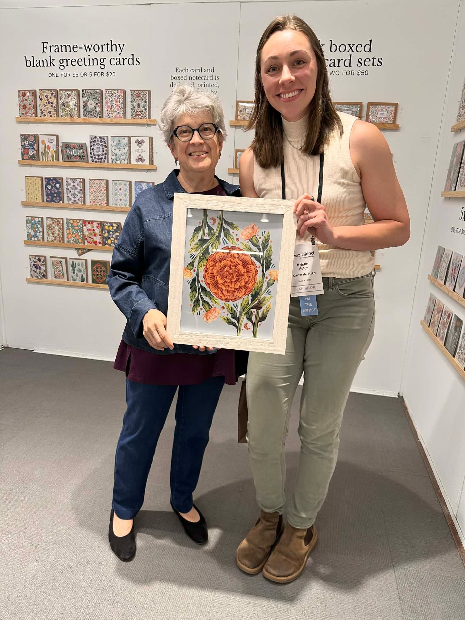 Two smiling people stand in a gallery, holding a framed floral artwork in front of walls lined with framed art cards.
