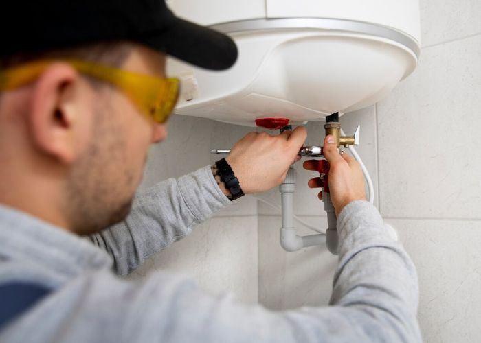 A technician is repairing or installing a water heater, connecting pipes and fittings under the unit.