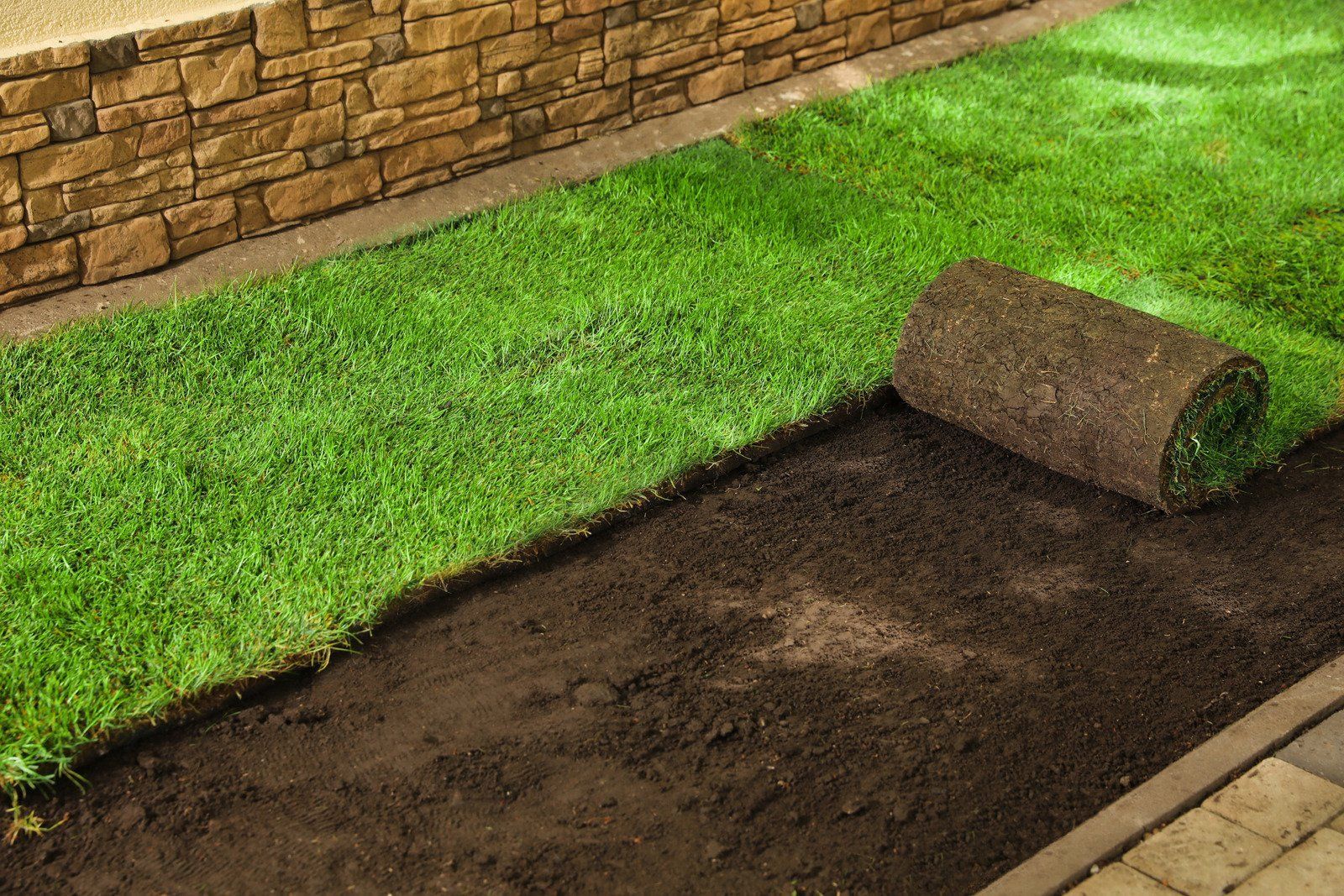 A partially installed lawn of green sod rolls next to dark, prepared soil beside a stone wall and paved walkway.