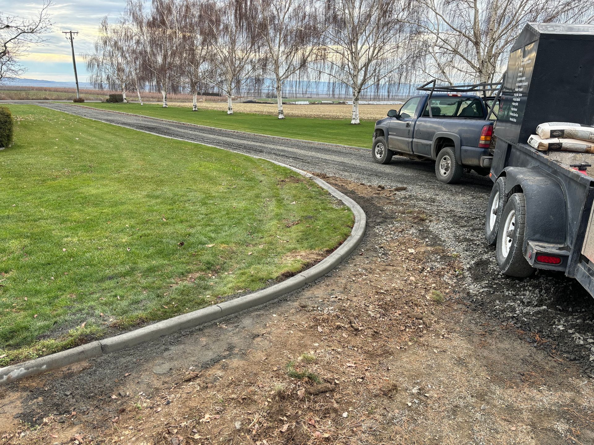A gray pickup truck with a trailer parked on a gravel driveway next to a curved concrete curb and a grassy lawn.