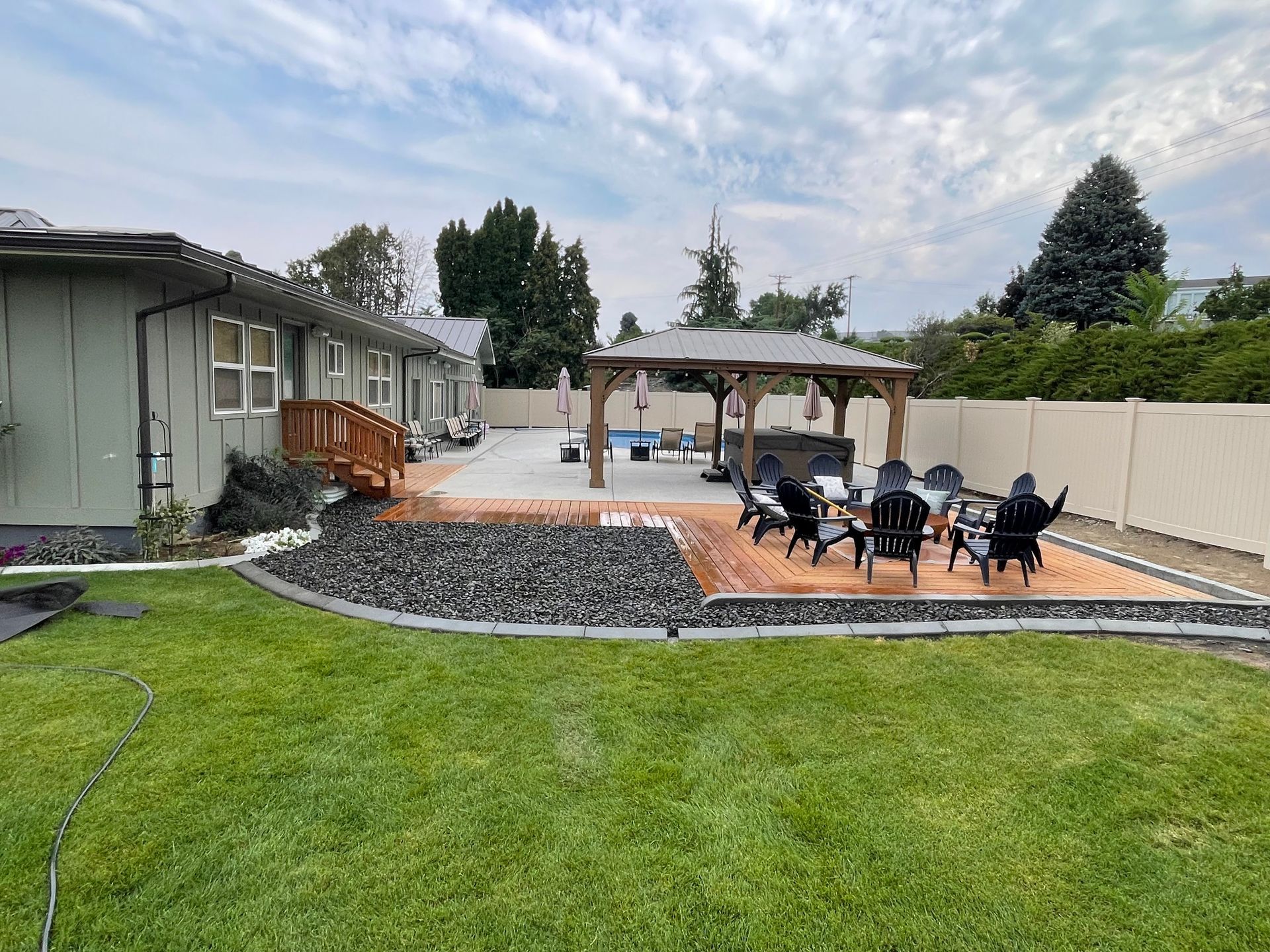 A backyard scene with a green lawn, a dark gravel border, a patio with dark chairs under a gazebo, and a light-colored house.